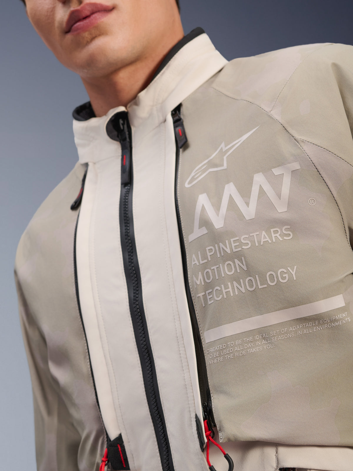 Chaqueta Alpinestars AMT-10LAB Drystar® XF, White Sand Aluminum Camo, primer plano del pecho con el logotipo de AMT Alpinestars Motion Technology, cremalleras impermeables con tiradores rojos y tejido textil texturizado resistente para aventura.