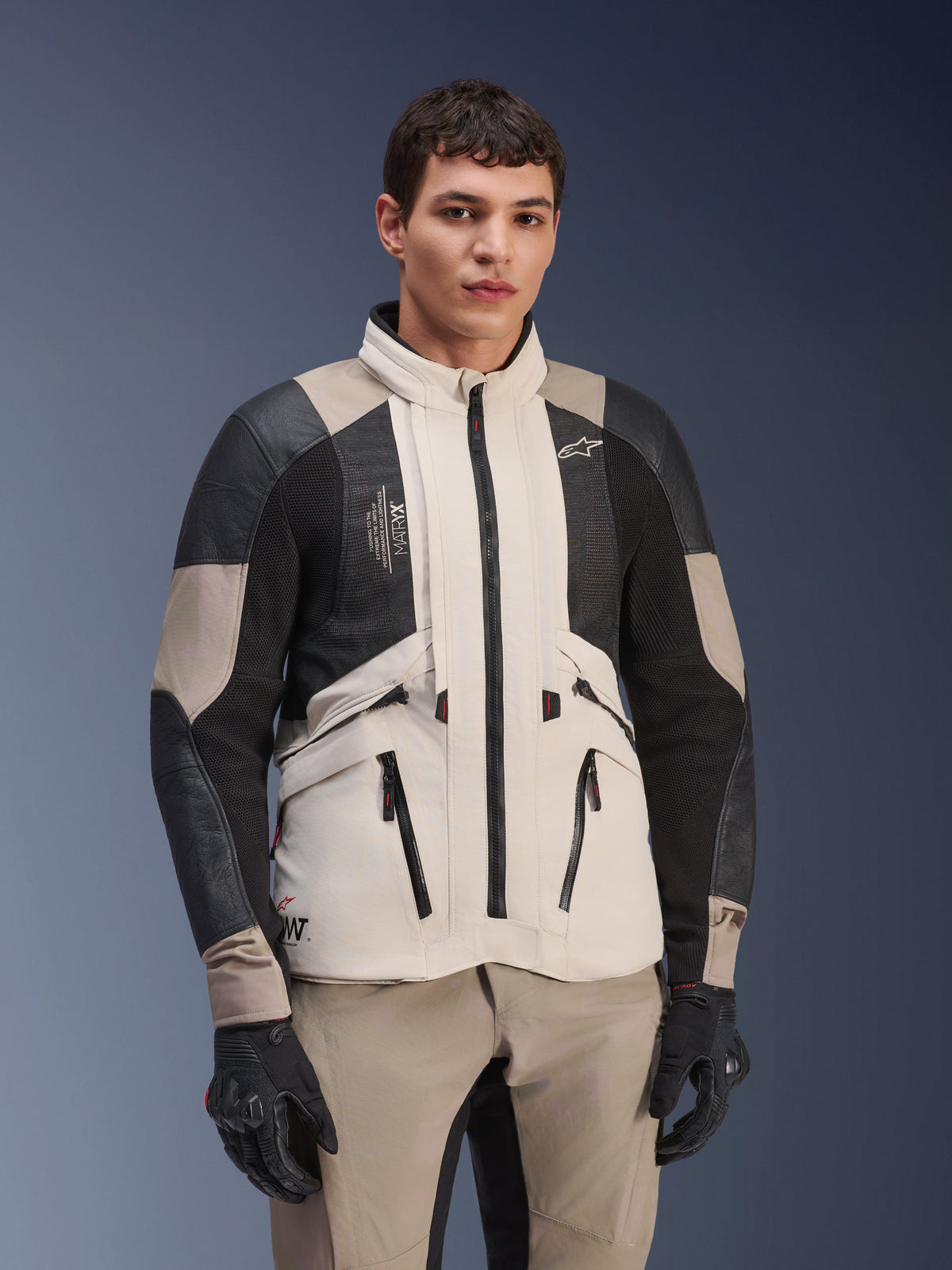 Chaqueta Alpinestars AMT-10LAB Drystar® XF, White Sand Aluminum Camo, chaqueta de aventura para motocicleta con paneles de malla MATRYX® negros y refuerzos de cuero, vestida por un modelo con guantes negros y pantalones color arena a juego, retrato de estudio
