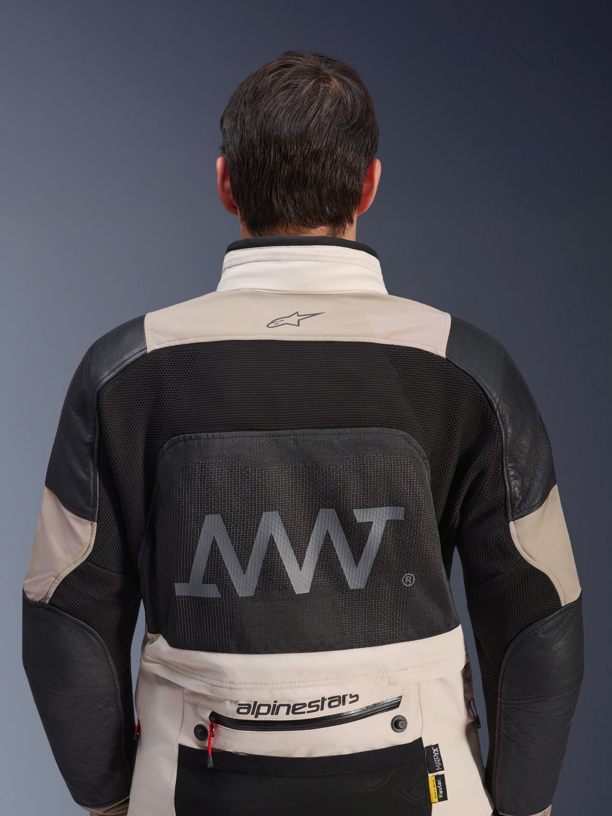Vista trasera de la chaqueta Alpinestars AMT-10LAB Drystar® XF en color arena blanca y negro, con un gran logotipo AMT en el panel de malla de la espalda, marca Alpinestars en la parte inferior de la espalda y construcción textil técnica para motociclismo de aventura