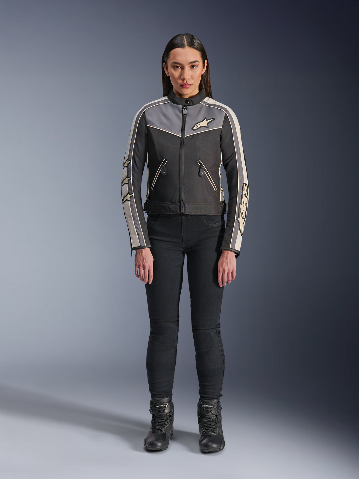 Mujer con la chaqueta Alpinestars Stella T-Dyno WR, negro, gris oscuro y crudo, vista frontal, chaqueta textil de moto con diseño de inspiración vintage, con logotipos en el pecho y las mangas, usada con pantalones negros y botas