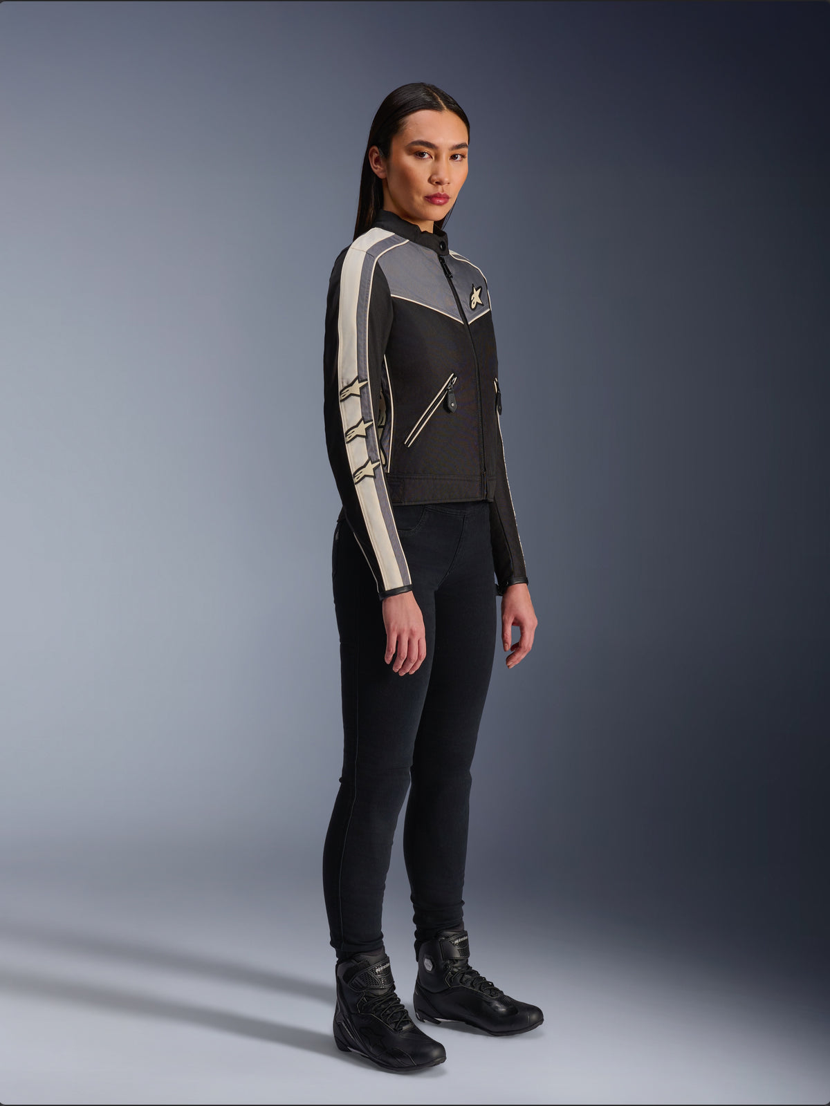 Alpinestars Stella T-Dyno WR Jacket, chaqueta de moto textil para mujer, en color negro, gris oscuro y crudo, con diseño de inspiración vintage, franjas crema en las mangas y logotipos de estrellas, combinada con pantalones negros y botas de moto, equipamiento de conducción urbana de estilo clásico