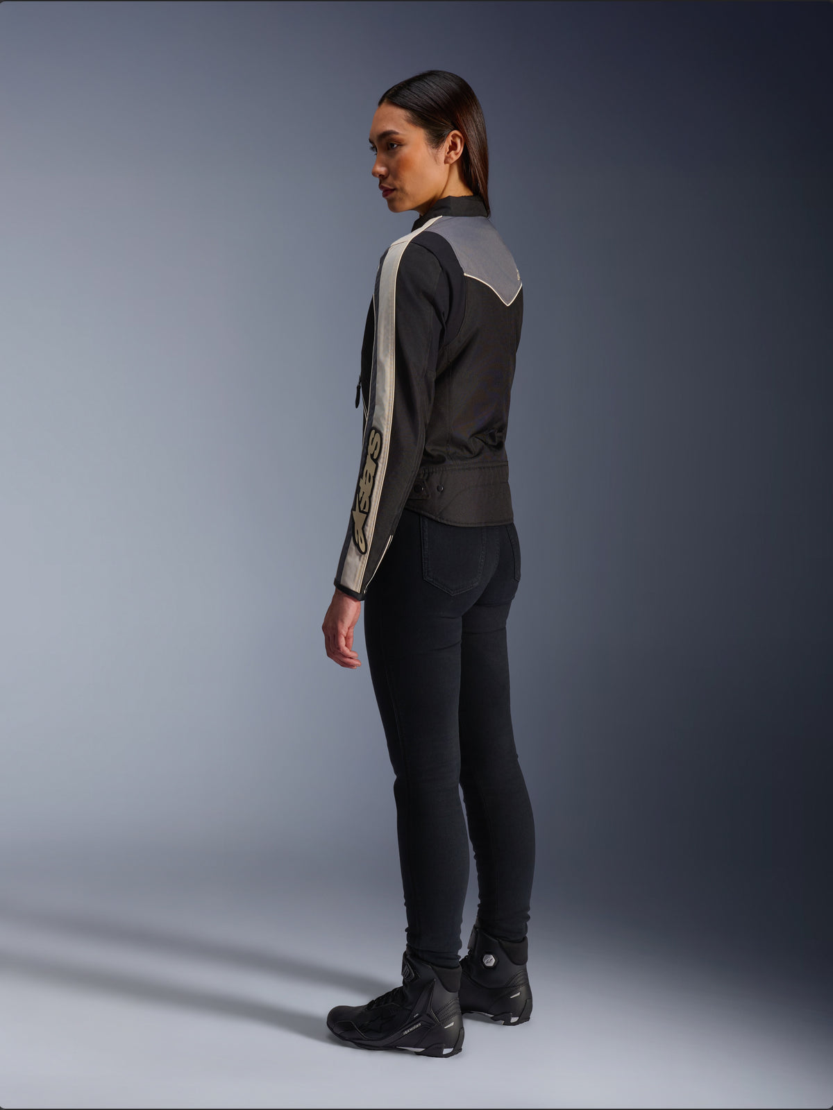 Modelo femenina vista de lado y desde atrás luciendo la chaqueta Alpinestars Stella T-Dyno WR en color Black Dark Gray Ecru, con un diseño de inspiración vintage con franjas color crudo en las mangas y un panel trasero gris, combinada con pantalones negros y calzado de conducción negro.