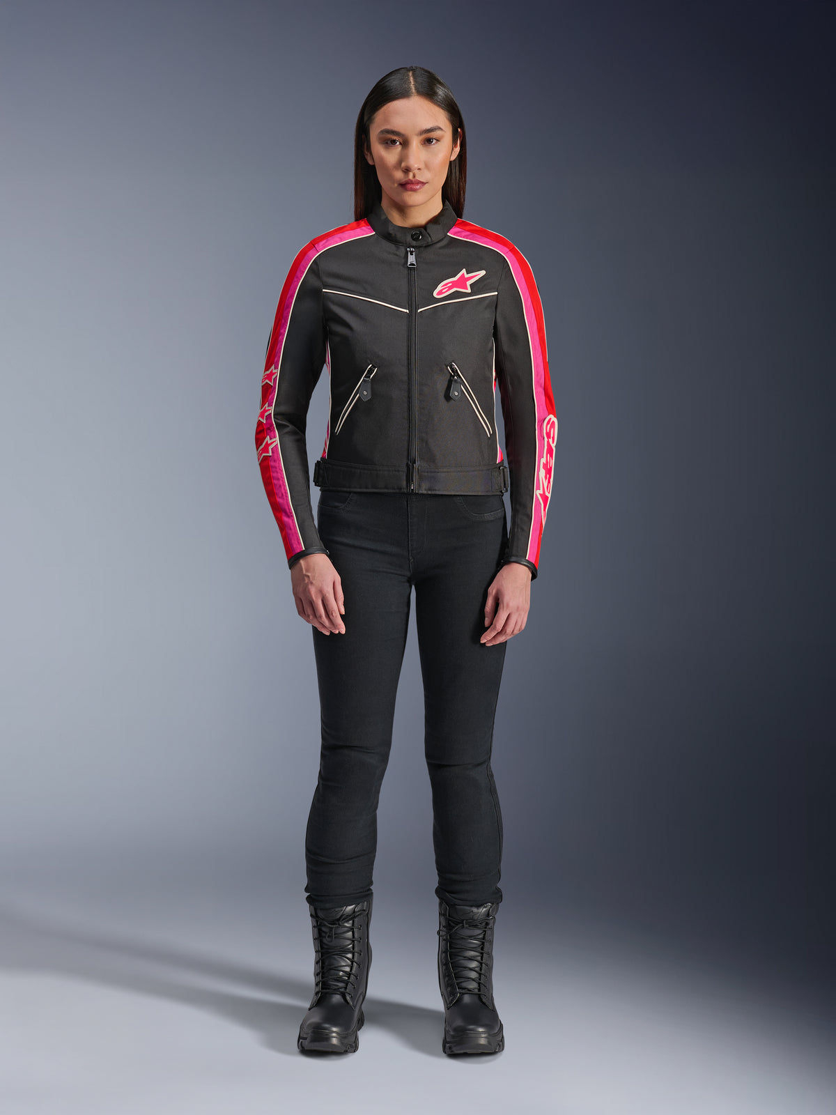 Mujer con la chaqueta Alpinestars Stella T-Dyno WR, negra con detalles en rojo brillante y fucsia, chaqueta textil de estilo vintage con logo rosa y franjas en las mangas, combinada con pantalones negros y botas con cordones, equipamiento urbano para motocicleta