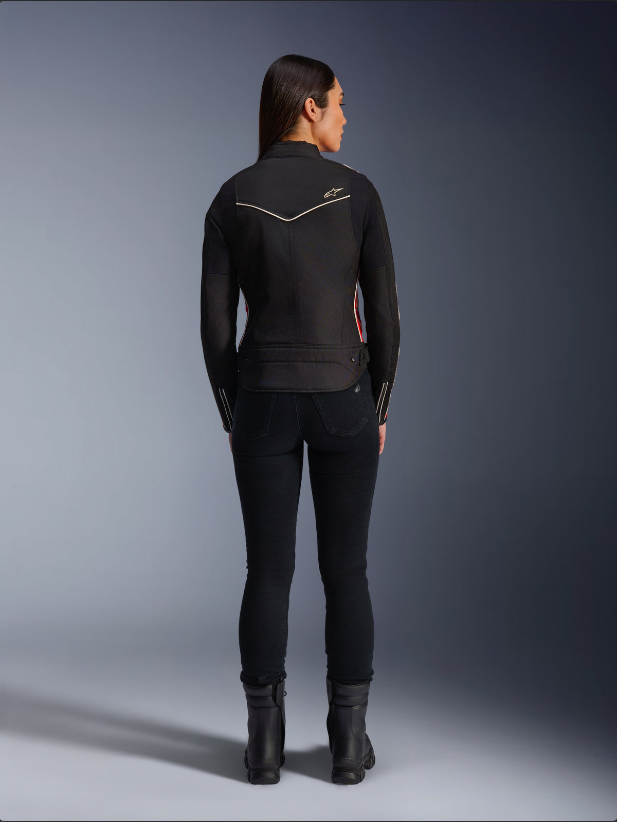 Modelo femenina de espaldas vistiendo la chaqueta Alpinestars Stella T-Dyno WR en color Black Bright Red Fuchsia, chaqueta textil de motociclismo con un ajuste de motociclista más corto, vista posterior que muestra el logotipo de la estrella blanca, combinada con pantalones negros y botas negras
