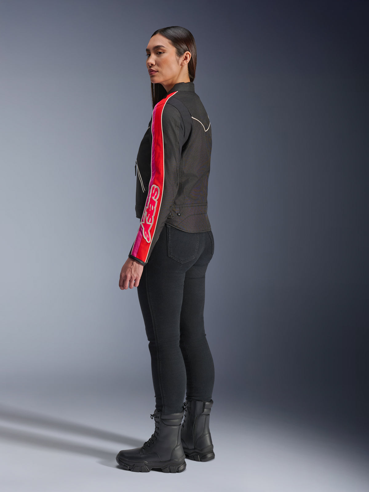 Modelo femenina vistiendo la chaqueta Alpinestars Stella T-Dyno WR en color negro, rojo brillante y fucsia, con llamativas franjas rojas y rosas y el logotipo de Alpinestars en la manga, combinada con pantalones de conducción negros y botas de moto negras en un entorno de estudio.