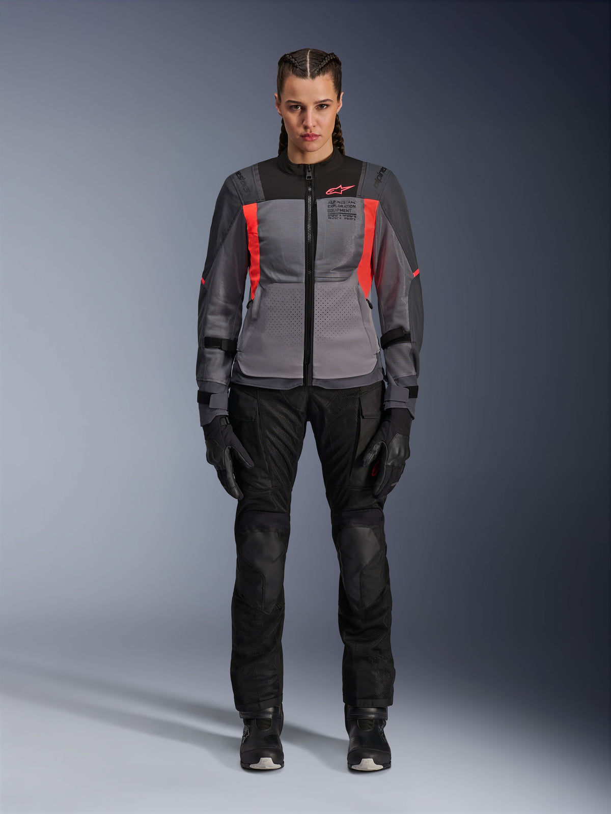 Mujer vistiendo la chaqueta Alpinestars Stella ST-2 Air en gris profundo, gris oscuro y coral flúor, combinada con pantalones y guantes de moto negros, vista frontal, equipamiento moderno de malla para turismo de aventura para mujer