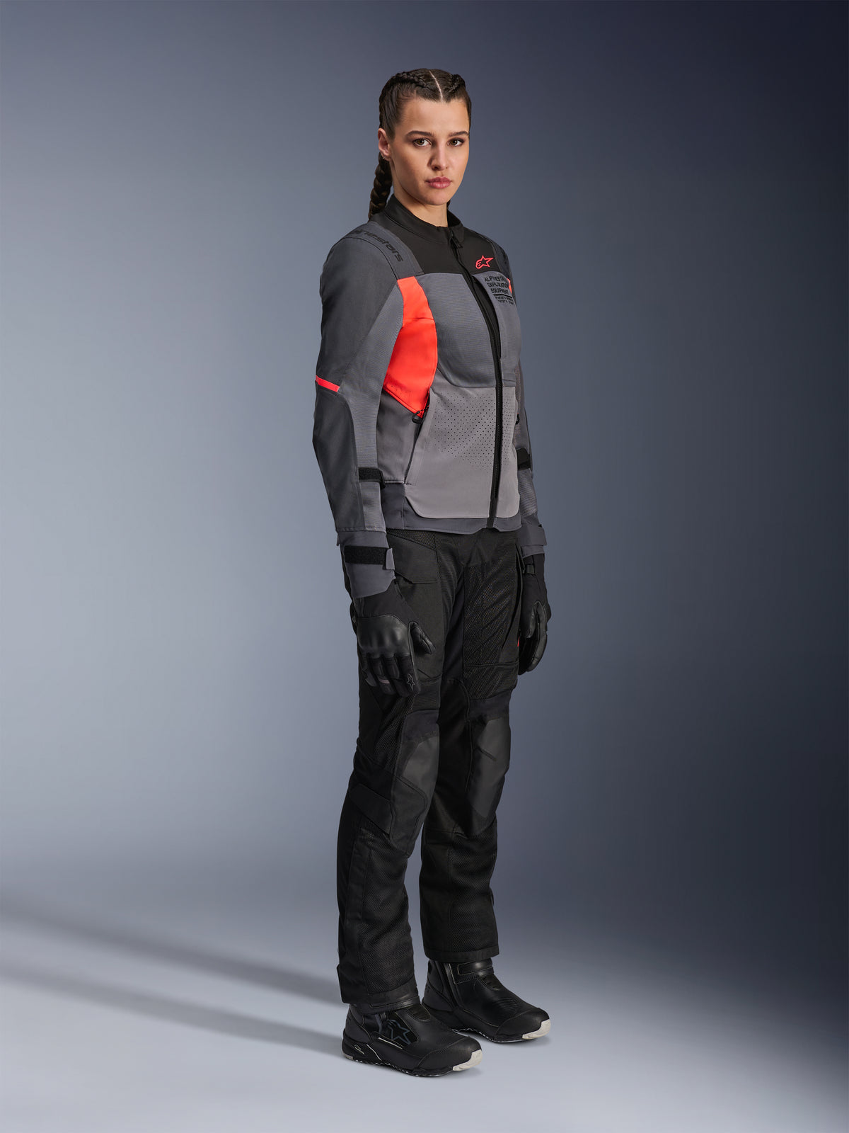 Motociclista con la chaqueta Alpinestars Stella ST-2 Air en gris intenso, gris oscuro y coral flúor, combinada con pantalones, guantes y botas de moto negros, con un diseño touring moderno con paneles de malla y detalles reflectantes.
