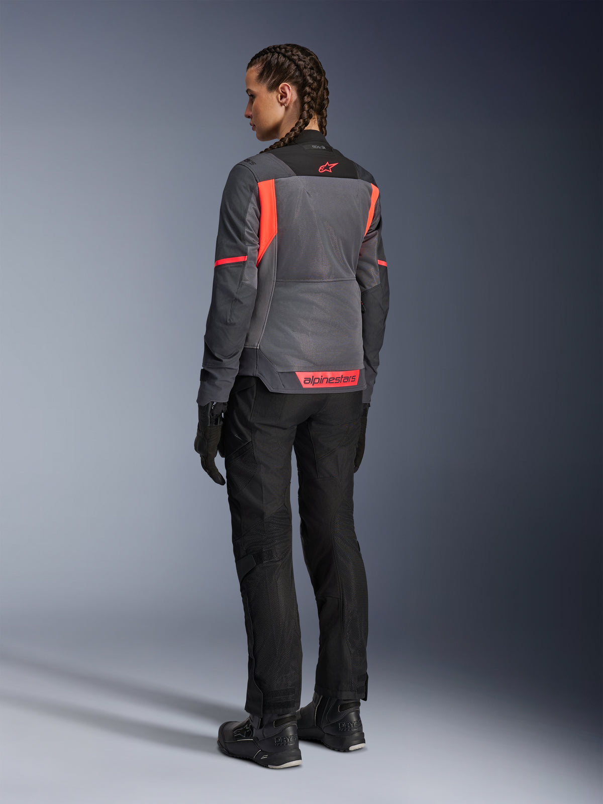 Mujer con la chaqueta Alpinestars Stella St-2 Air, chaqueta de touring para moto, en gris profundo y gris oscuro con acentos en coral flúor, combinada con pantalones y guantes de conducción negros, vista trasera que muestra grandes paneles de malla y marca reflectante en coral, diseñada para aventuras de verano