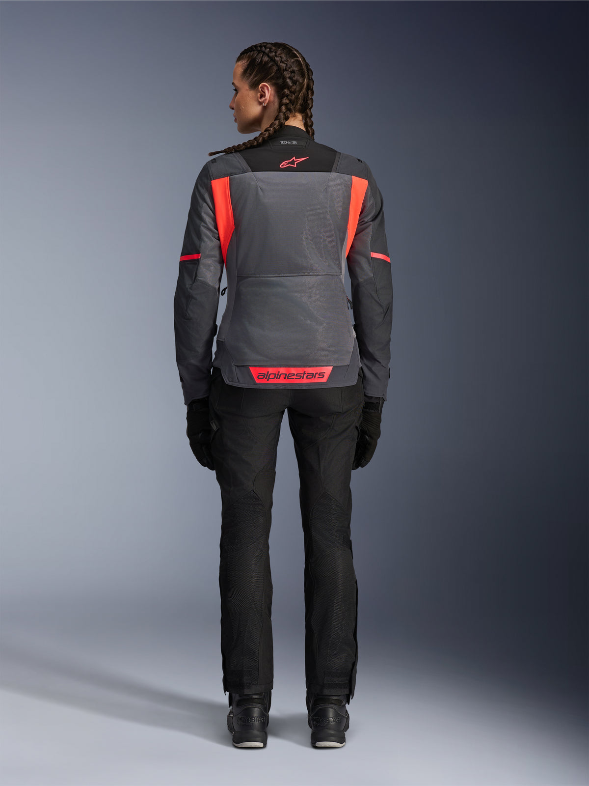 Vista trasera de una modelo con la chaqueta Alpinestars Stella St-2 Air para mujer en Deep Gray y Dark Gray con detalles en Fluo Coral, que presenta grandes paneles de ventilación de malla en la espalda y detalles de logotipos reflectantes, combinada con pantalones de moto y guantes negros.