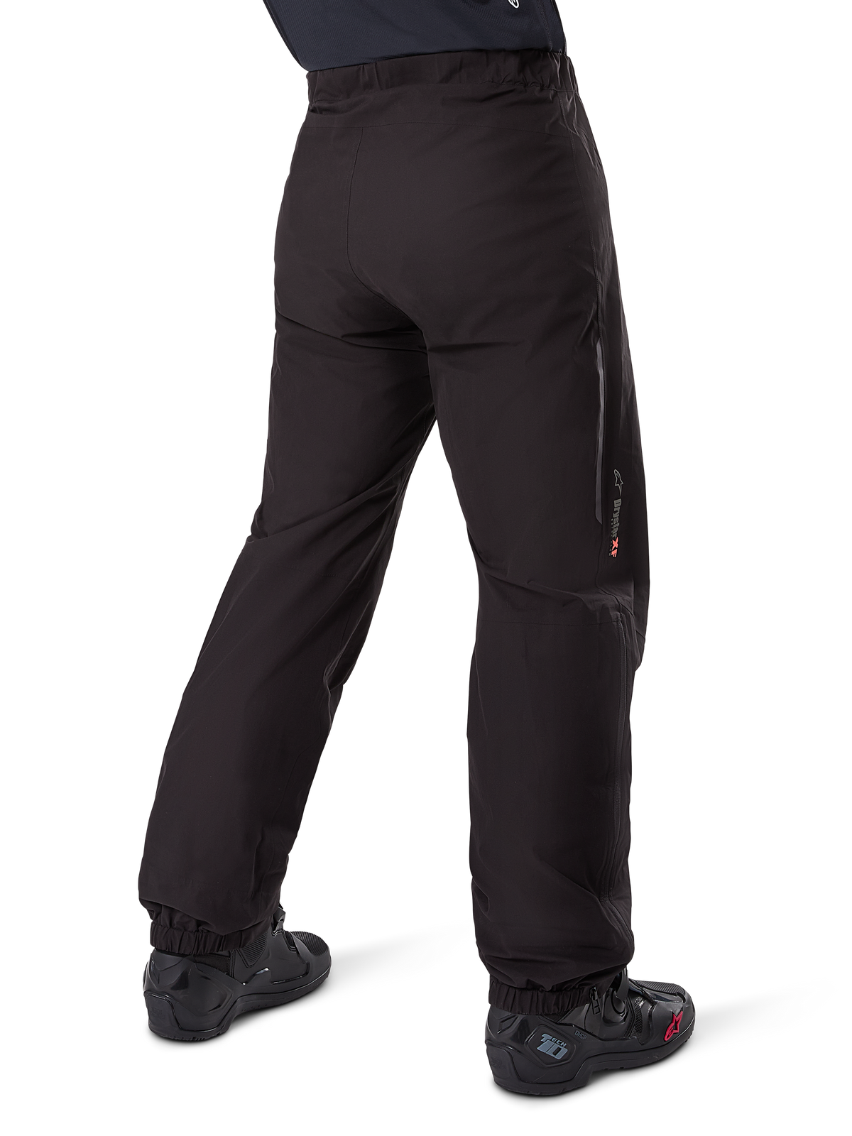 Forro de pantalón Alpinestars Amt-8 Stretch Drystar® XF, negro, vista posterior que muestra el forro impermeable sobre botas de moto negras, con la marca Drystar XF en la parte inferior de la pierna, puños elásticos en los tobillos y material elástico ligero para turismo de aventura.