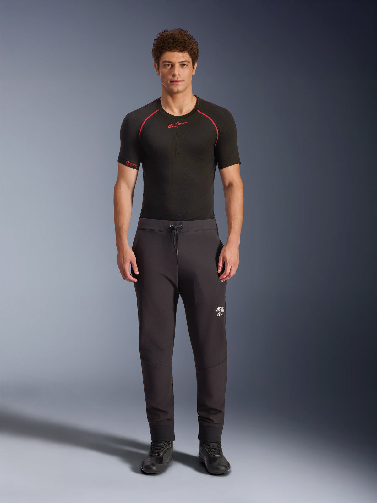 Alpinestars Aeroshell Pants, Black motorcycle softshell pants, worn with a black short-sleeved base layer and black shoes, featuring an elastic drawstring waist, white Astars logo on the thigh, and elastic cuffs, modern athletic silhouette for road riding