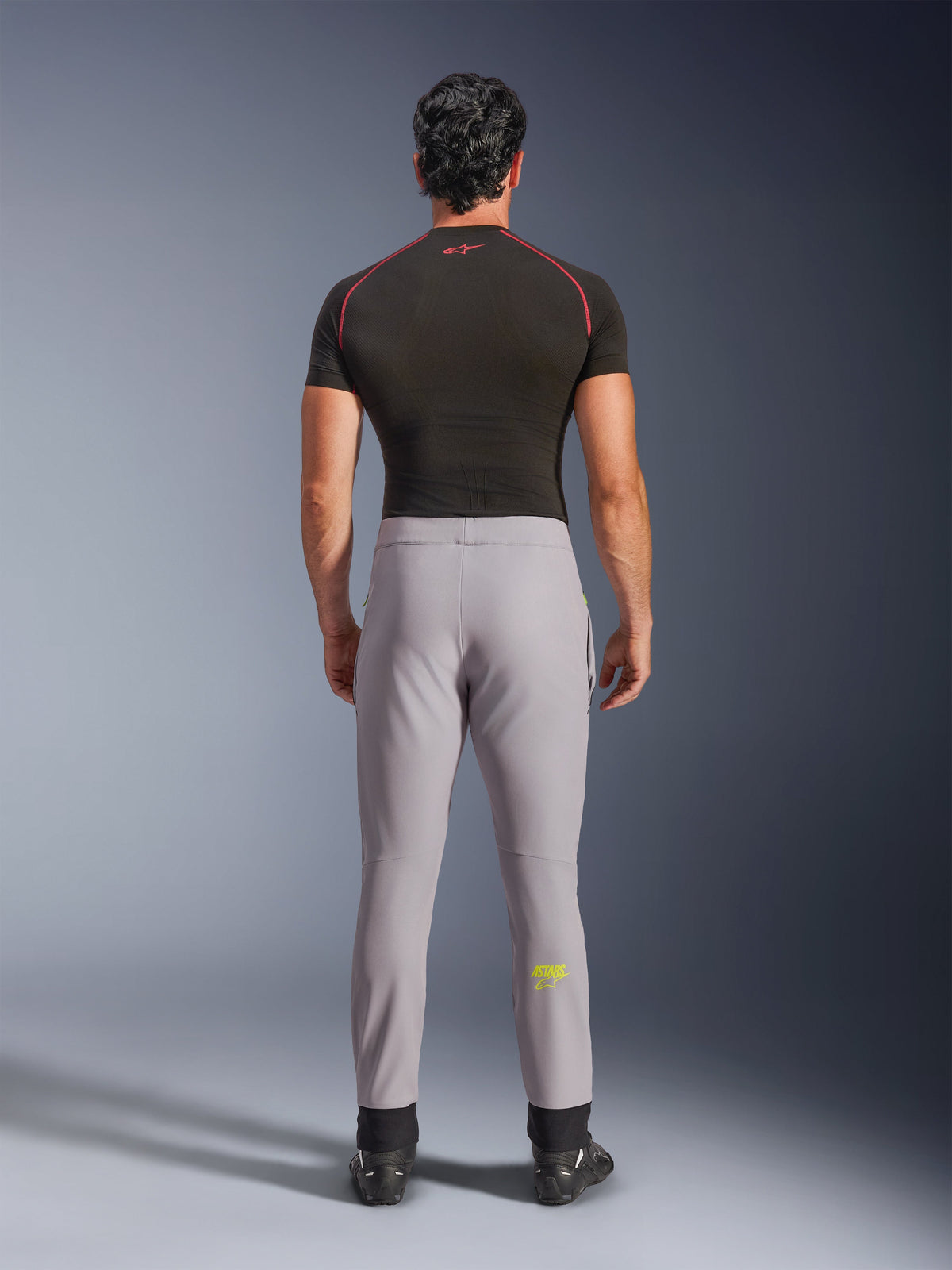 Alpinestars Aeroshell Pants, pantalones de moto softshell deportivos crossover, gris medio con logotipo amarillo flúor, vista trasera, usados con camiseta de compresión negra y calzado de conducción, con cintura elástica y silueta entallada