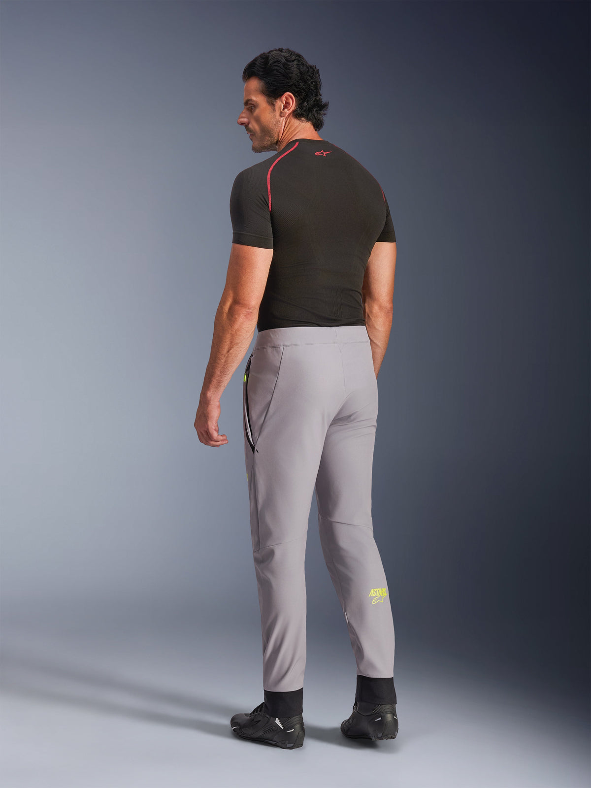 Pantalones Alpinestars Aeroshell, gris medio amarillo flúor, vista trasera de modelo masculino con pantalones de moto softshell elásticos, logotipo Astars amarillo en la pantorrilla izquierda, combinados con una capa base negra y calzado de conducción para una silueta deportiva moderna