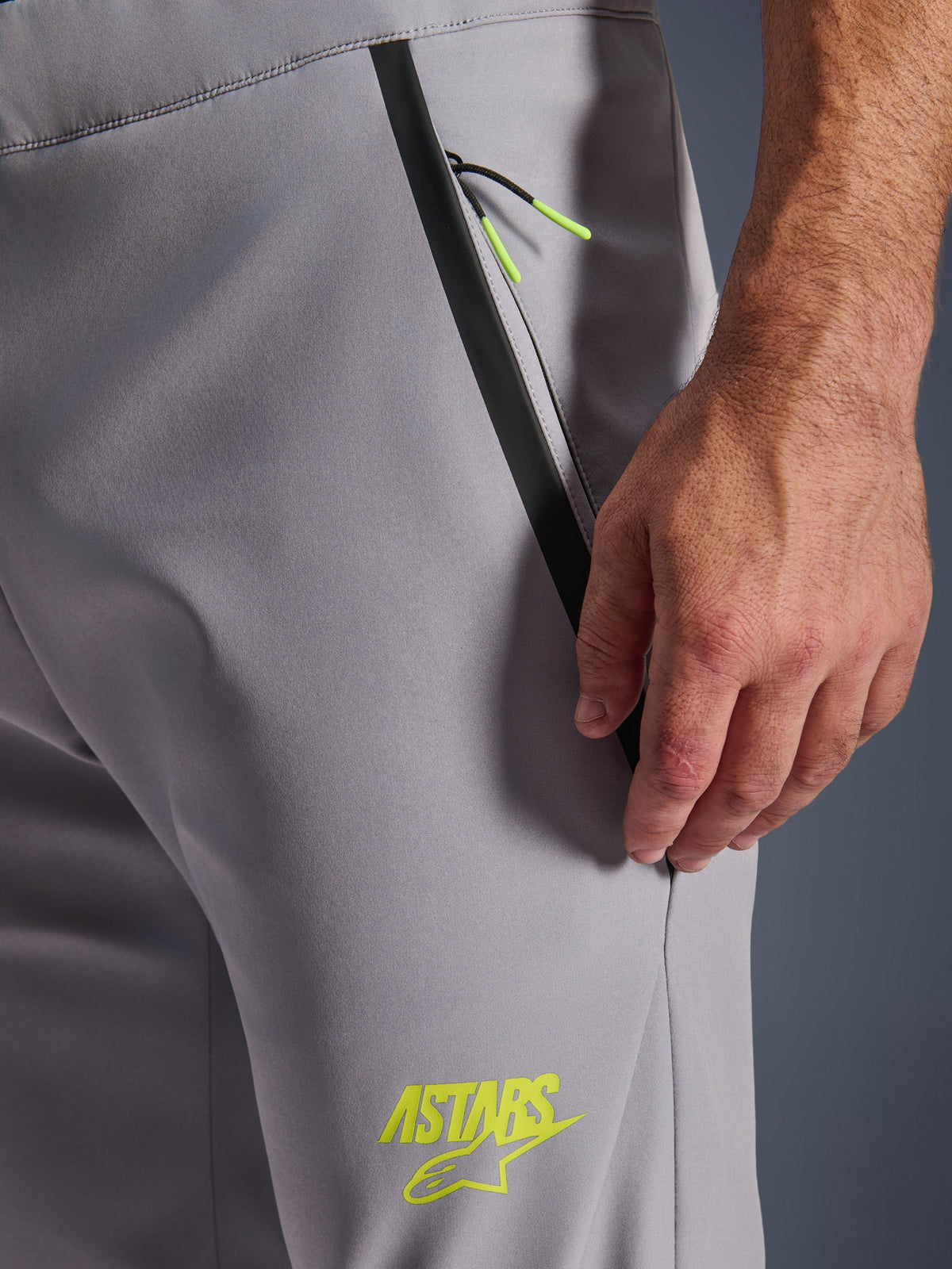 Pantalones Alpinestars Aeroshell, gris medio con detalles en amarillo flúor, primer plano del bolsillo frontal con cremallera con ribete negro y tiradores amarillos, logotipo ASTARS amarillo en el muslo, material softshell elástico para motociclismo