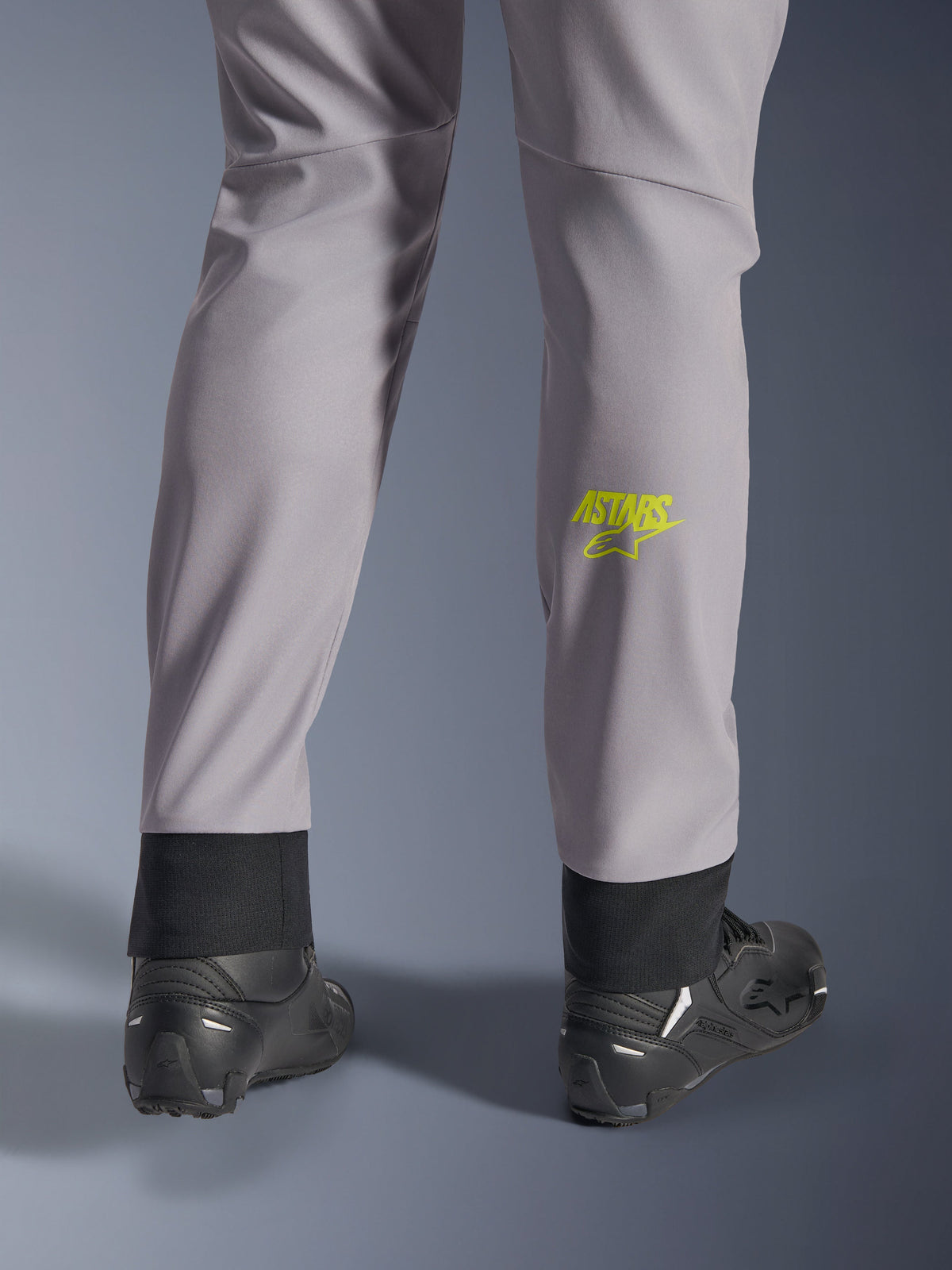 Pantalones Alpinestars Aeroshell, gris medio con logo Astars en amarillo flúor en la pantorrilla, vistos desde atrás con calzado de moto negro, con dobladillo inferior elástico y silueta cónica moderna