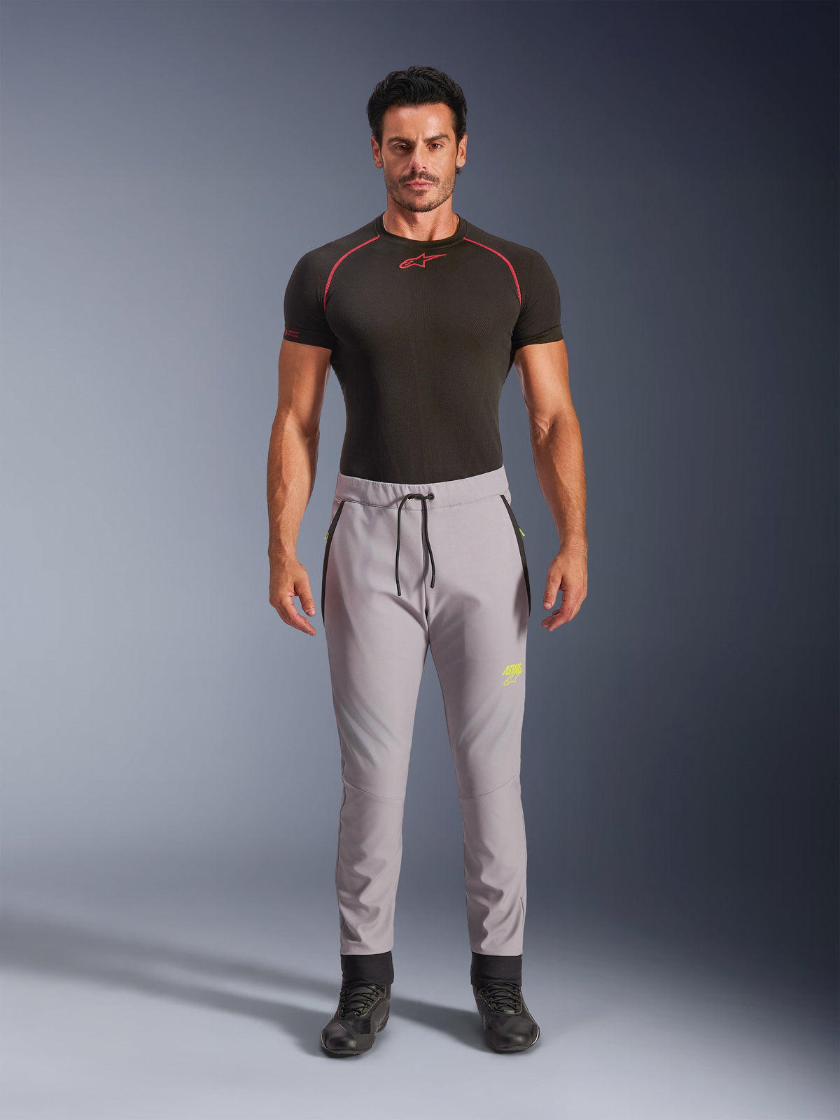 Modelo masculino con pantalones Alpinestars Aeroshell en gris medio y amarillo flúor, pantalones softshell deportivos crossover con cintura elástica con cordón y bolsillos con cremallera, combinados con una camiseta técnica de manga corta negra, diseño atlético moderno para conducción urbana y en carretera