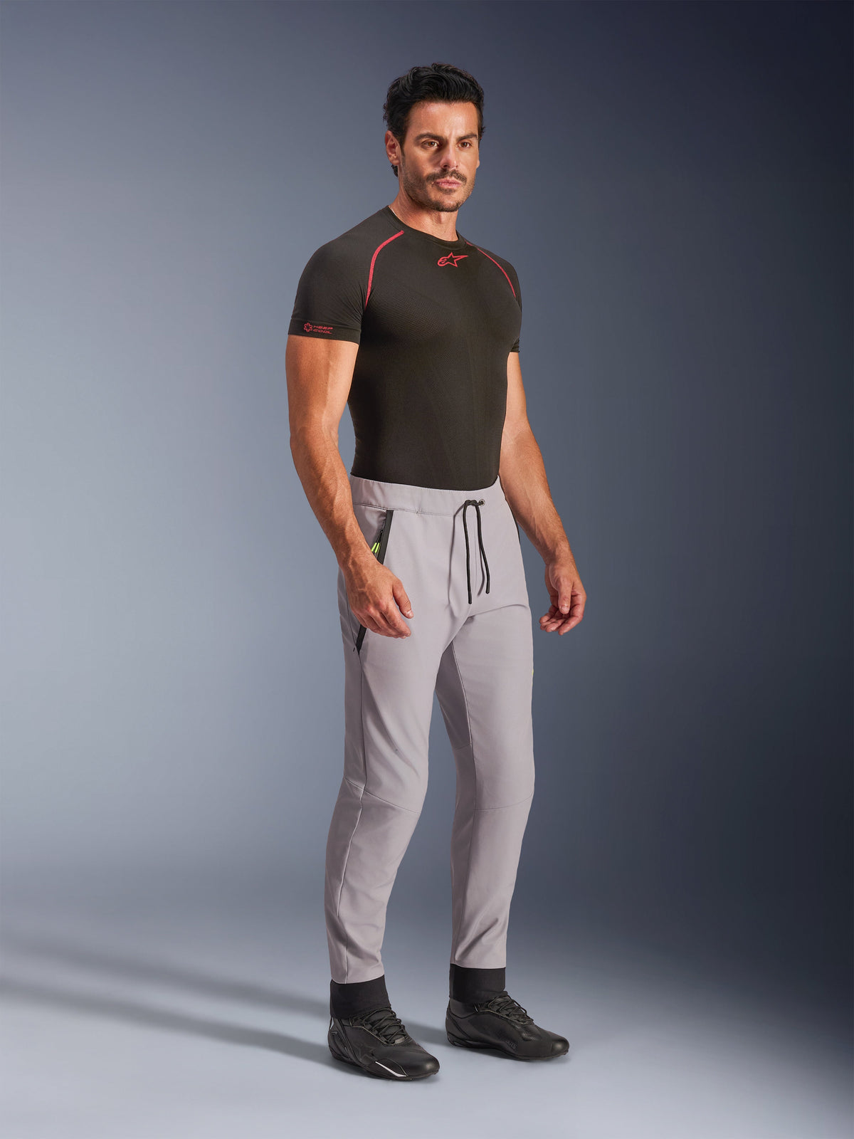 Pantalones Alpinestars Aeroshell, Gris Medio con detalles en Amarillo Flúor, pantalones softshell crossover usados por un modelo con una capa base negra y calzado negro, con un corte moderno entallado, cintura elástica con cordón negro y bolsillos con cremallera.