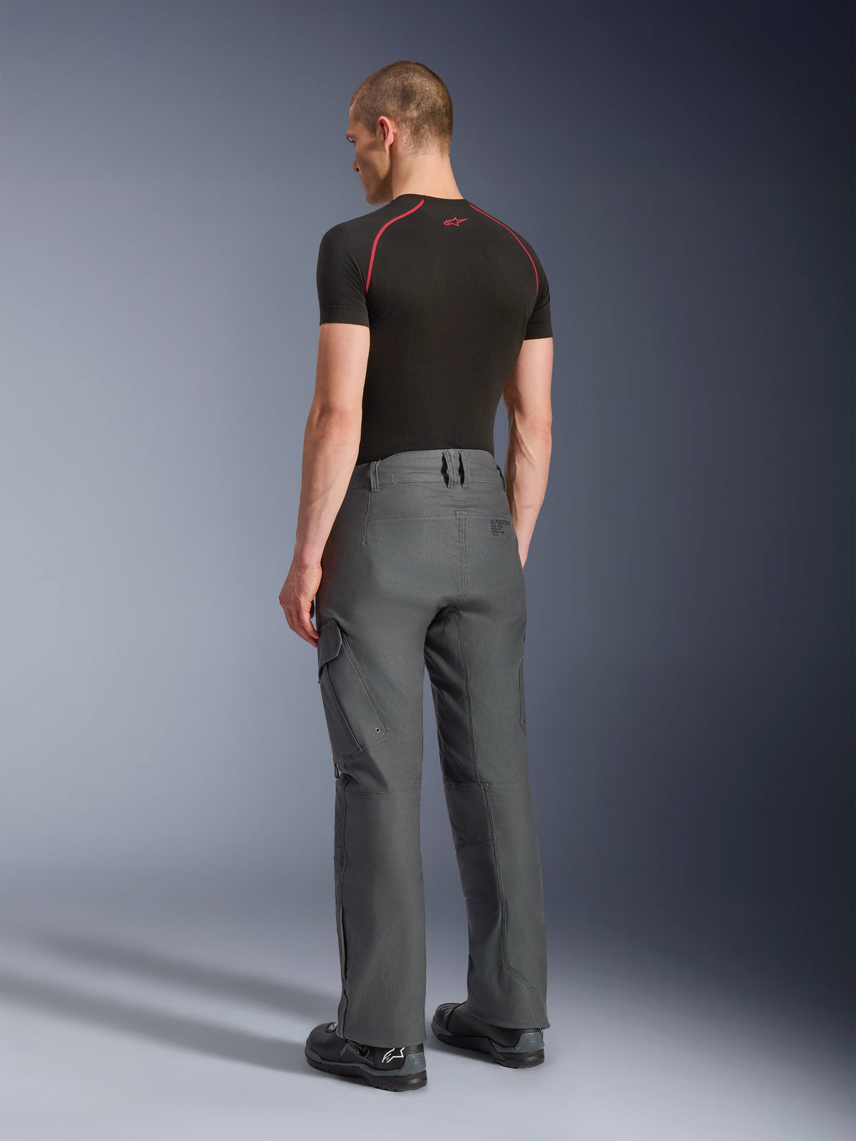 Pantalones Alpinestars Borrego Drystar®, pantalones de moto de aventura, gris antracita, vista trasera, con aspecto de lona encerada y bolsillos de carga, combinados con una camiseta negra y botas de moto, diseño resistente para carretera y adventure touring
