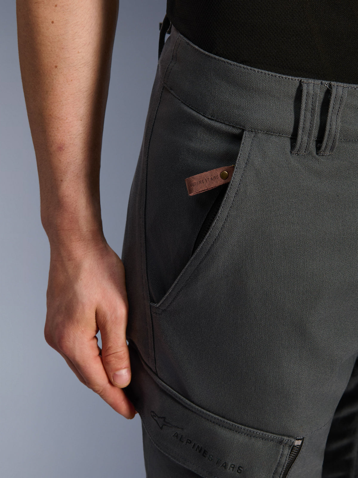 Pantalones Alpinestars Borrego Drystar®, pantalones de moto de aventura, gris antracita, detalle de primer plano del bolsillo y la cadera, textura de lona encerada con una etiqueta de logo de cuero marrón y la marca Alpinestars bordada en el bolsillo de carga del muslo.