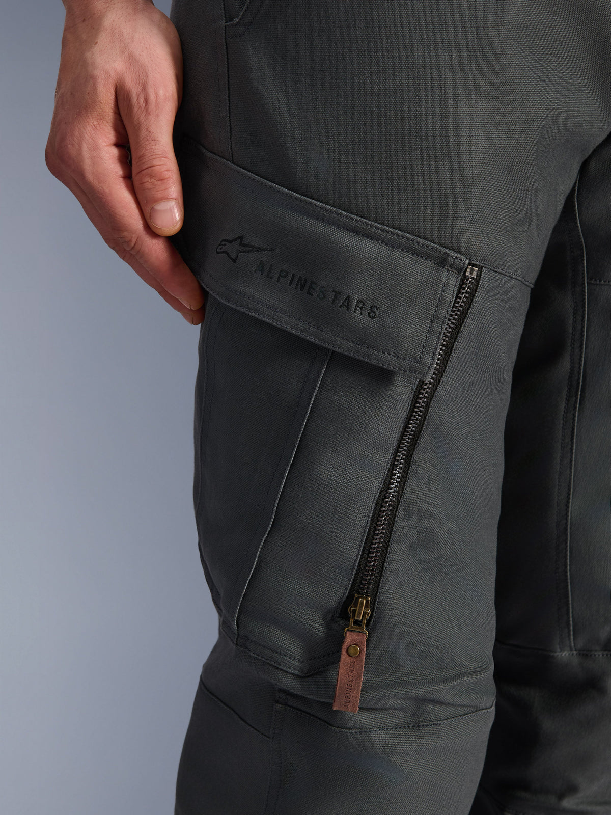 Pantalones Alpinestars Borrego Drystar®, gris antracita, primer plano del bolsillo cargo en el muslo con marca bordada, material de lona encerada texturizada, cremallera de ventilación vertical con tirador de cuero marrón