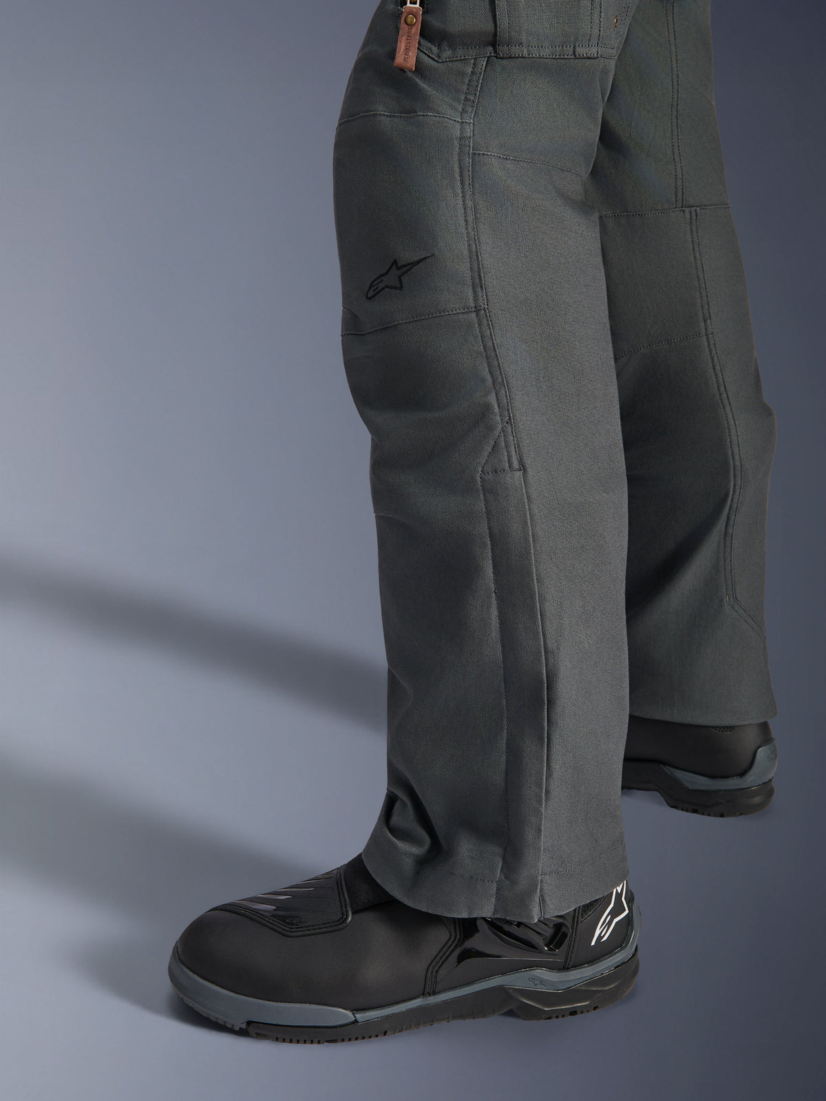 Pantalones Alpinestars Borrego Drystar®, pantalones de moto de aventura gris antracita con apariencia de lona encerada, construcción textil mostrada de lado con botas de moto negras, diseño robusto para carretera y touring de aventura