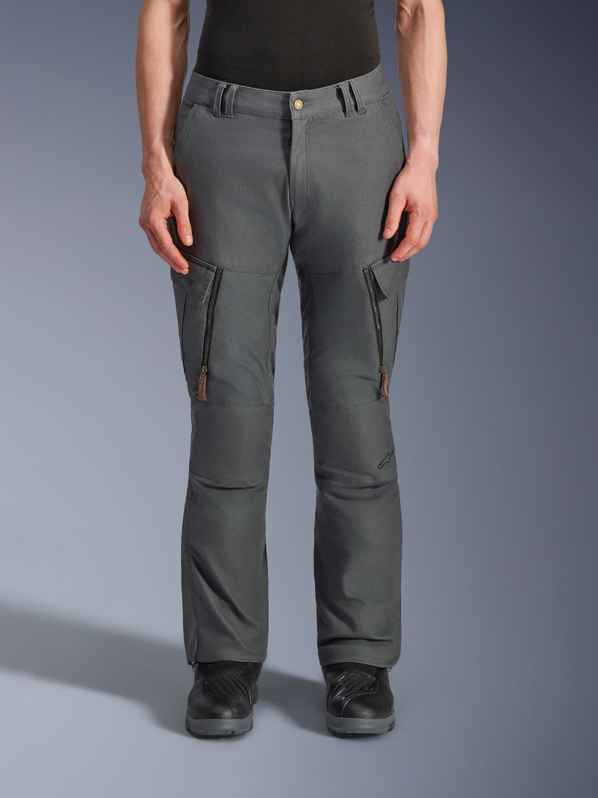Pantalones Alpinestars Borrego Drystar®, pantalones de aventura para moto, gris antracita, con bolsillos verticales con cremallera en los muslos y tiradores de cuero, cierre de botón en la cintura y trabillas, usados por un modelo con botas de moto negras