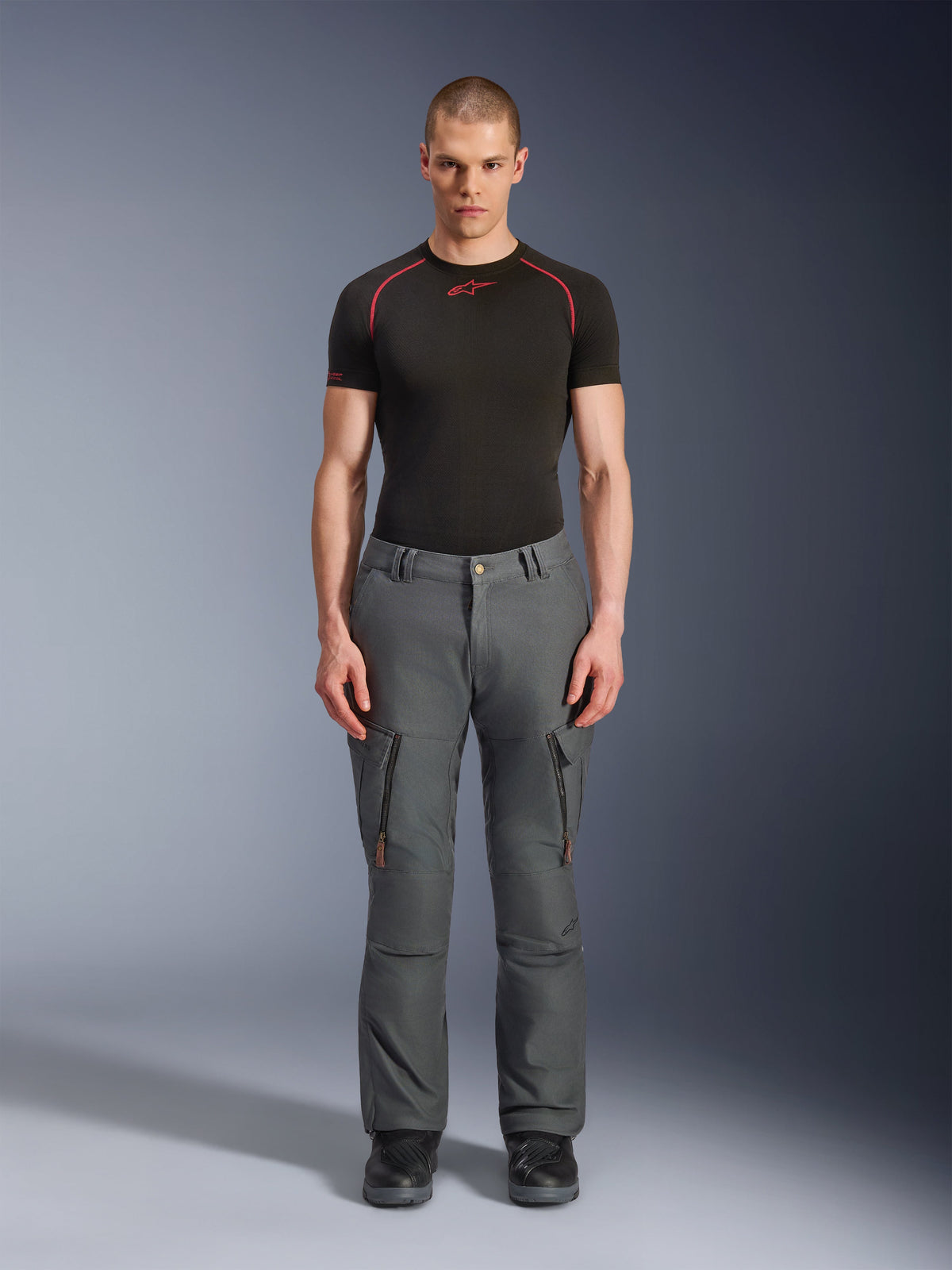 Pantalones Alpinestars Borrego Drystar®, pantalones de moto de aventura grises, vestidos por un modelo con una camiseta técnica negra y roja y botas negras, con bolsillos cargo con cremalleras verticales, apariencia de lona encerada resistente, diseño duradero para turismo de aventura y conducción en carretera