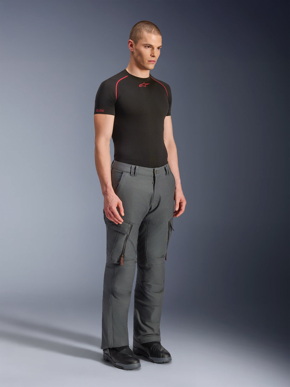 Modelo masculino vistiendo pantalones Alpinestars Borrego Drystar®, pantalones de moto de aventura en color gris antracita, diseño inspirado en ropa de trabajo con bolsillos cargo y ventilaciones con cremallera, combinados con una capa base negra de manga corta y botas negras