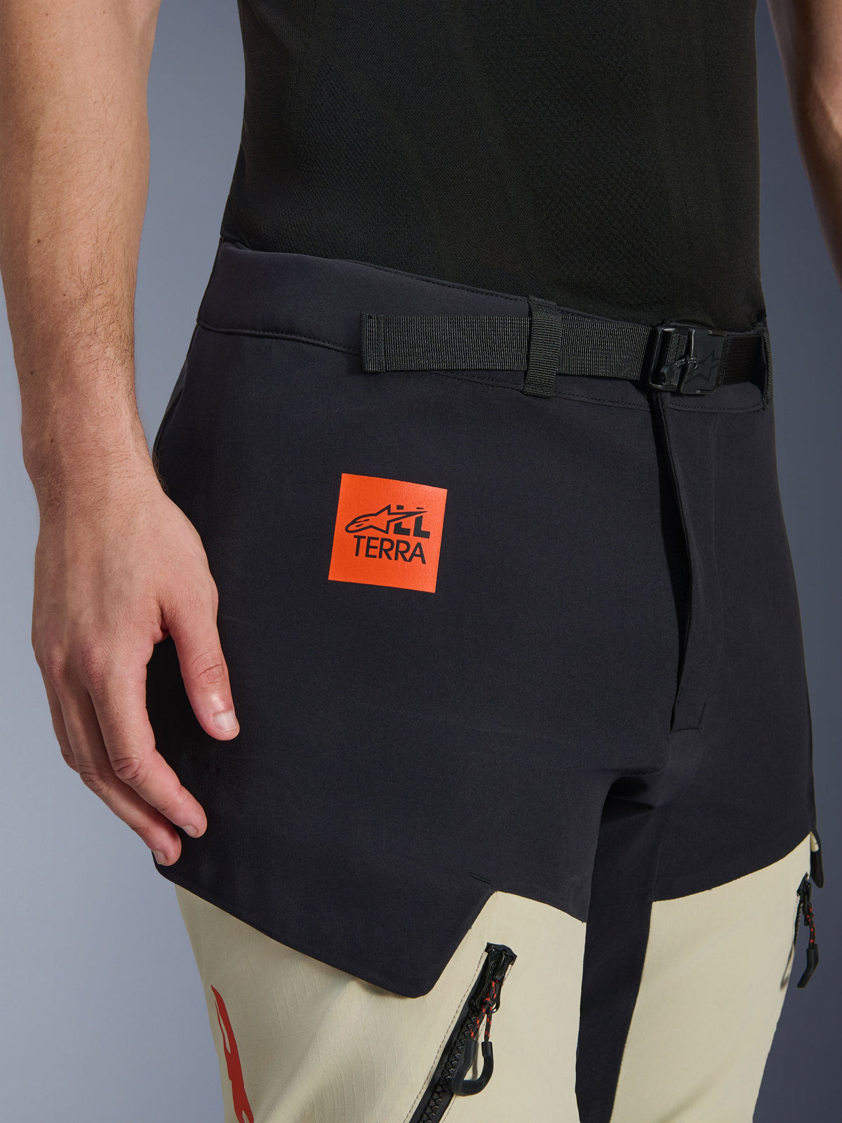 Pantalones Alpinestars Halo Pro Drystar® XF, pantalones laminados de moto de aventura, negro Mastic y beige, primer plano con el parche naranja del logo 'ALL TERRA', cinturón ajustable negro y tejido técnico resistente con cremalleras impermeables