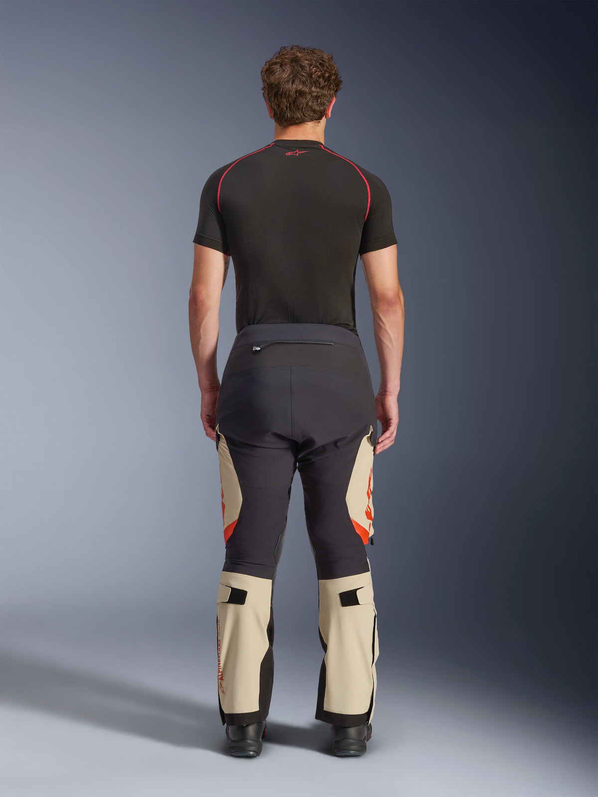 Pantalones Alpinestars Halo Pro Drystar® XF Laminados, Mastic Black, vista trasera en un modelo, bloques de color negro y beige con logotipos naranjas, pantalones de moto adventure touring con construcción textil duradera y secciones ajustables en la pantorrilla