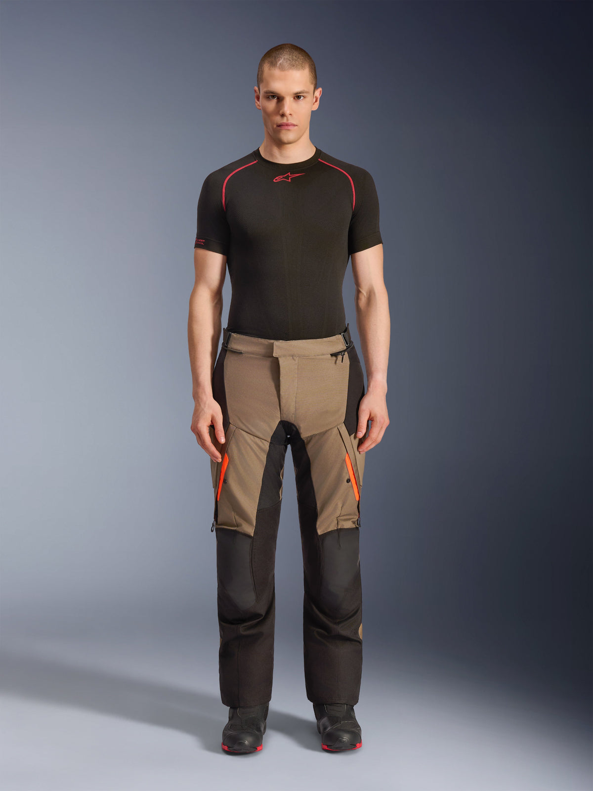 Modelo masculino luciendo los pantalones Alpinestars Andes V4 Drystar® en Walnut Black, pantalones de moto de aventura combinados con una capa base negra y botas, con detalles de cremalleras naranjas en los muslos y paneles negros reforzados para durabilidad en todo terreno.