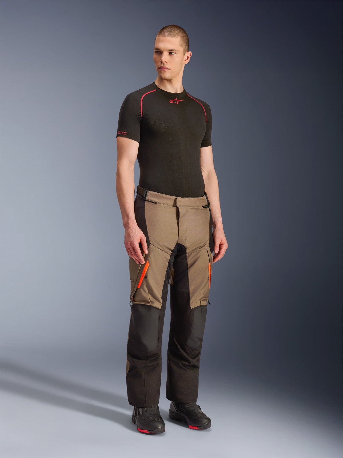 Pantalones Alpinestars Andes V4 Drystar®, pantalones de moto para Adventure Touring, color Walnut Black con detalles en naranja, usados por un modelo con una capa base negra de manga corta y botas negras, con paneles textiles reforzados y bolsillos de carga, diseño robusto para conducción en cualquier clima