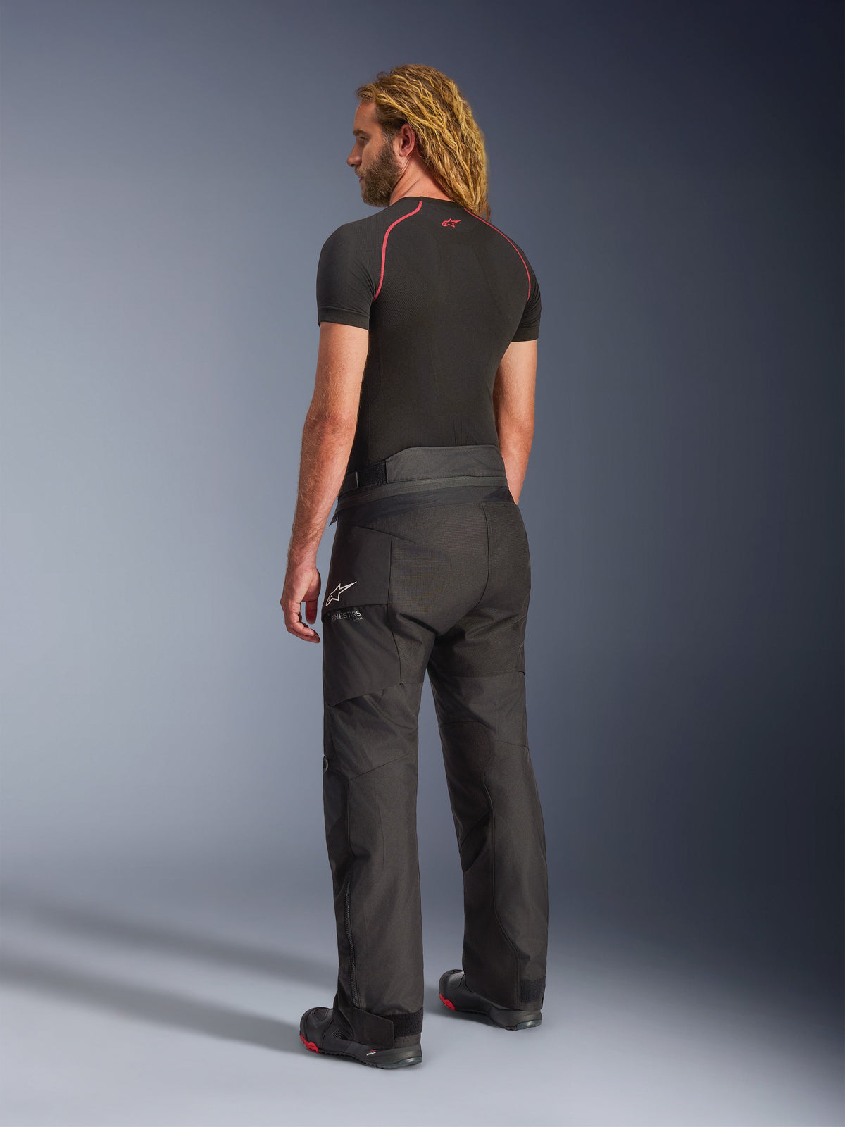 Pantalones Alpinestars Andes V4 Drystar®, pantalones de moto para aventura y touring en color negro, vistos desde una perspectiva trasera lateral en un modelo, con el logotipo blanco de Alpinestars en la cadera, combinados con una prenda base negra de manga corta y botas de moto, construcción textil resistente para carretera y aventura.
