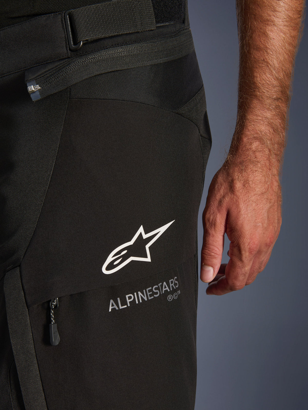 Detalle en primer plano de los pantalones Alpinestars Andes V4 Drystar® en color negro, con el logotipo y la marca Alpinestars en blanco en la solapa de un bolsillo de carga, con correa de ajuste de cintura y cremallera de conexión visibles, diseñados para el touring de aventura en moto.