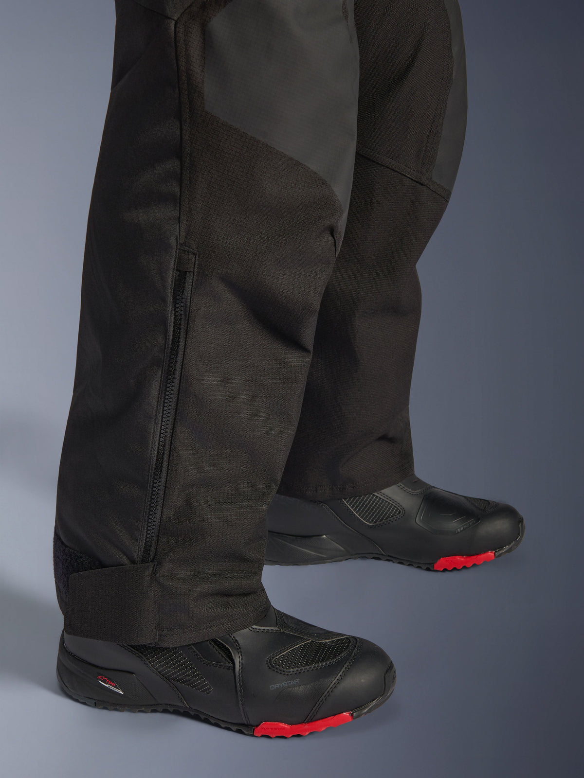 Pantalones Alpinestars Andes V4 Drystar®, pantalones de moto de aventura negros, primer plano de la parte inferior de la pierna con detalle de cremallera lateral, textil duradero con refuerzos de ripstop, combinados con botas negras con suelas rojas