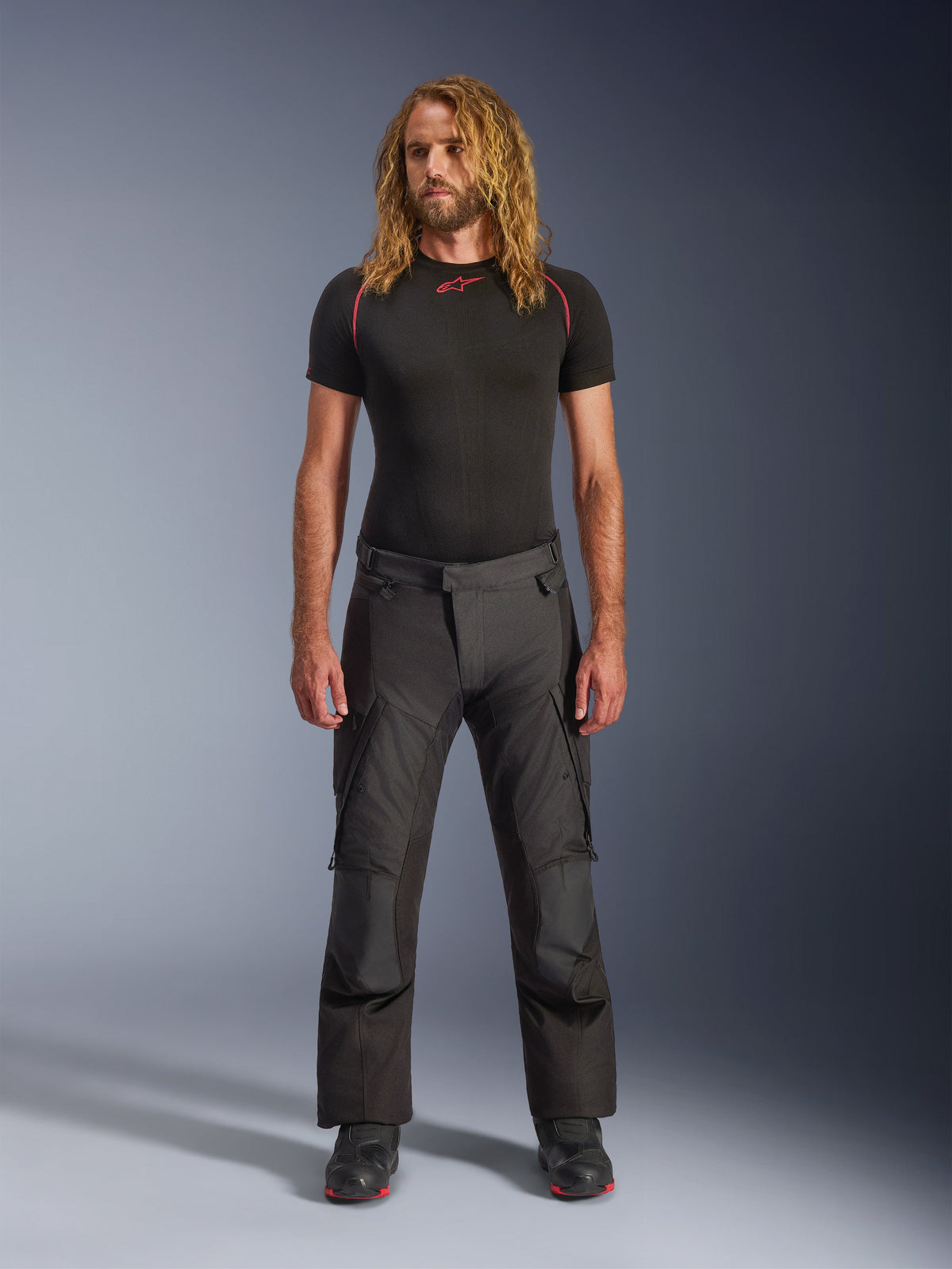 Pantalones Alpinestars Andes V4 Drystar®, pantalones negros de aventura para motociclismo, combinados con una capa base negra de manga corta y botas de conducción, con paneles reforzados y bolsillos tipo cargo, confección textil resistente para uso en cualquier clima