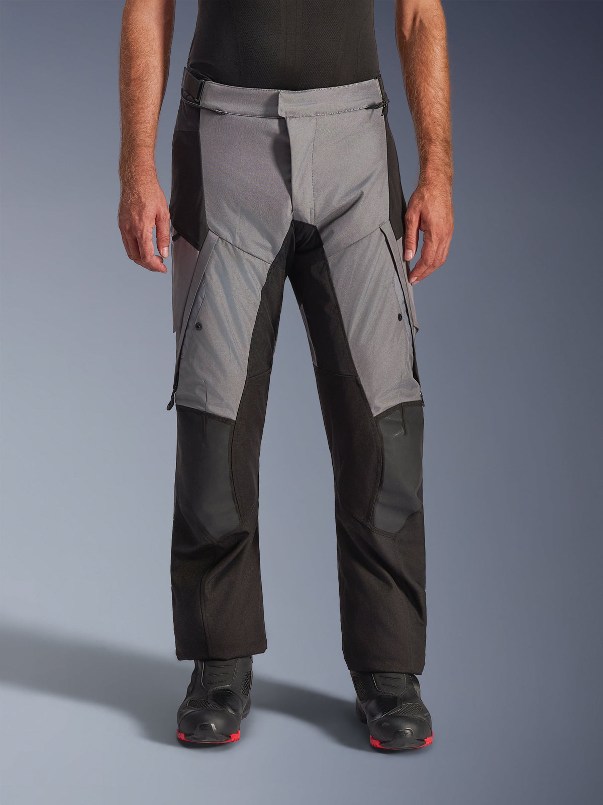 Pantalones Alpinestars Andes V4 Drystar®, pantalones de moto para turismo de aventura, gris y negro, vista frontal usados por un modelo con botas de motociclismo negras, con parches reforzados en las rodillas y bolsillos de carga en los muslos, construcción textil impermeable y transpirable para todo tipo de clima