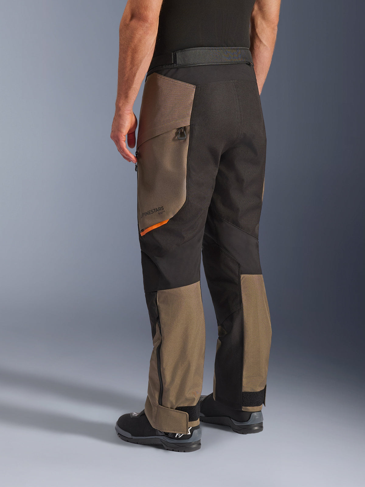 Alpinestars Cusco Drystar® Pants, pantalones de motociclismo de aventura, color Walnut Black con un pequeño detalle naranja, vista trasera de tres cuartos que muestra la construcción textil y paneles color nogal, usados con botas de motociclista negras.