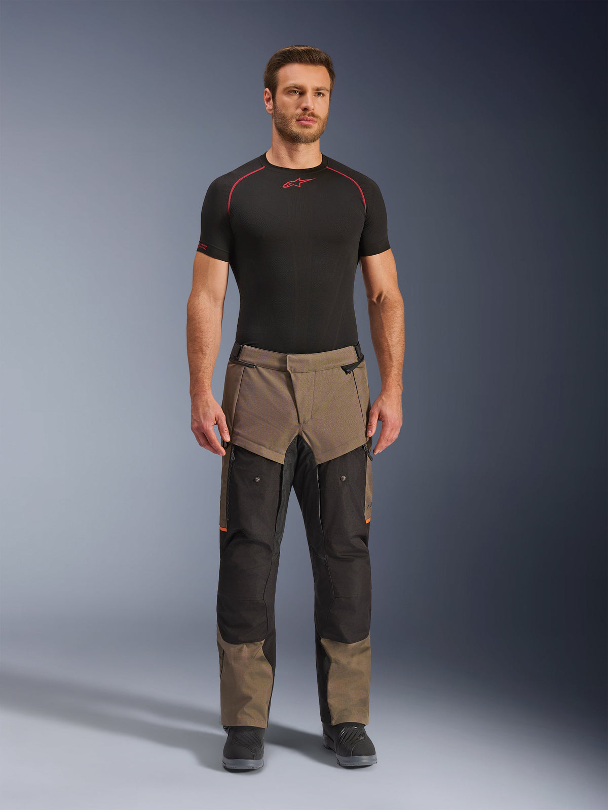 Pantalones Alpinestars Cusco Drystar®, Walnut Black, pantalones de motociclismo adventure touring, lucidos por un modelo masculino con una prenda de base negra y botas negras, con paneles en color nogal y negro, detalles en naranja y cremalleras de ventilación directa en los muslos.