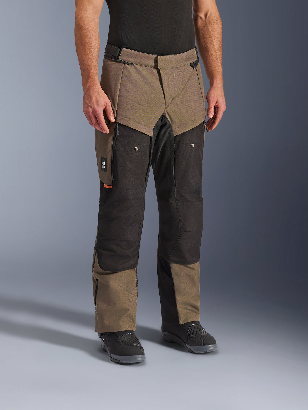 Pantalones Alpinestars Cusco Drystar®, pantalones de motociclismo adventure, color Walnut Black, mostrados en un modelo con botas negras, con un diseño de múltiples paneles en marrón y negro, bolsillos con cremallera y rodillas reforzadas para touring