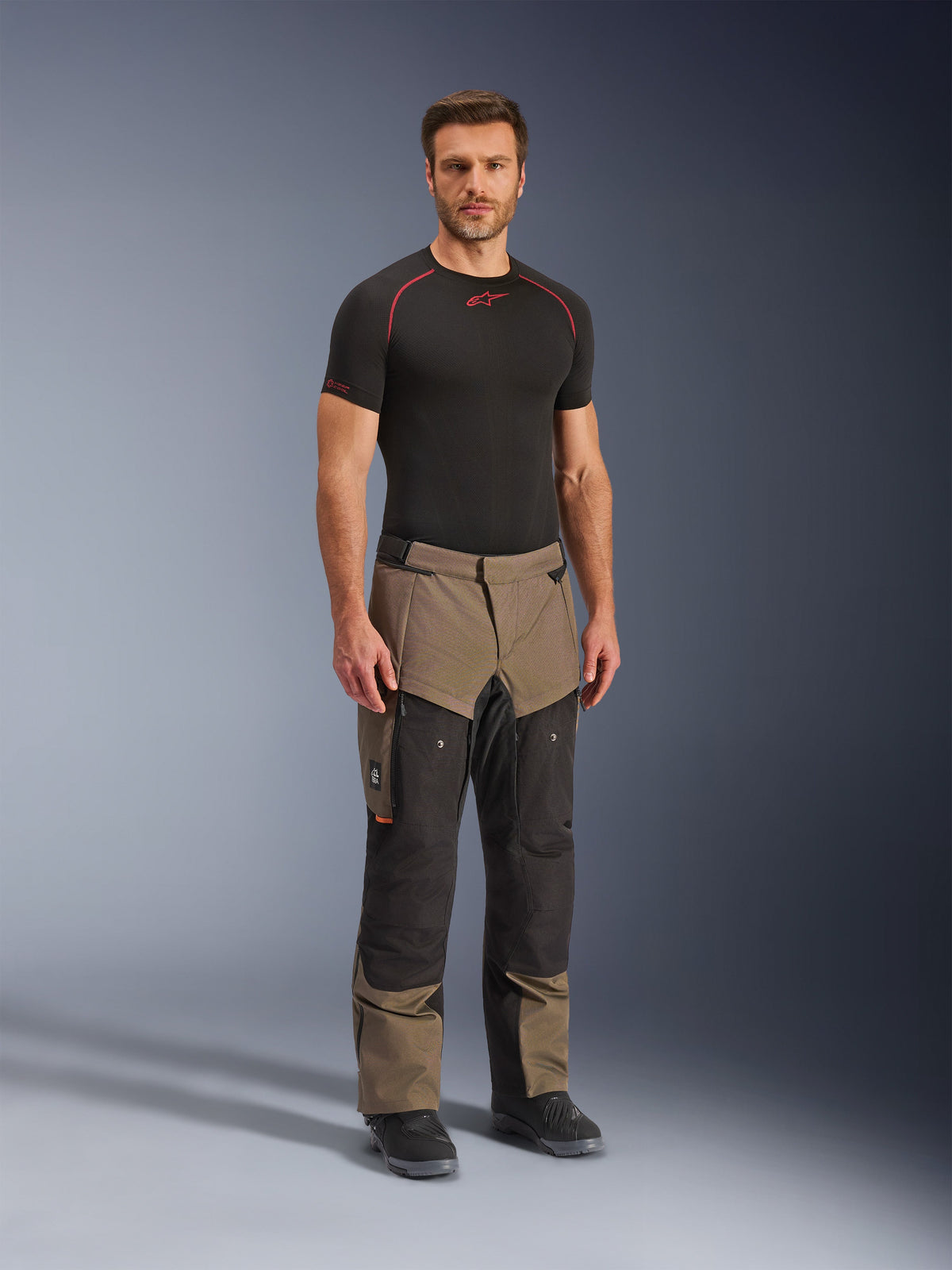 Pantalones Alpinestars Cusco Drystar®, pantalones de motociclismo adventure, color nogal y negro, usados por un modelo con una capa base negra y botas, con grandes bolsillos cargo en los muslos, cremalleras de ventilación y un diseño robusto bitono.
