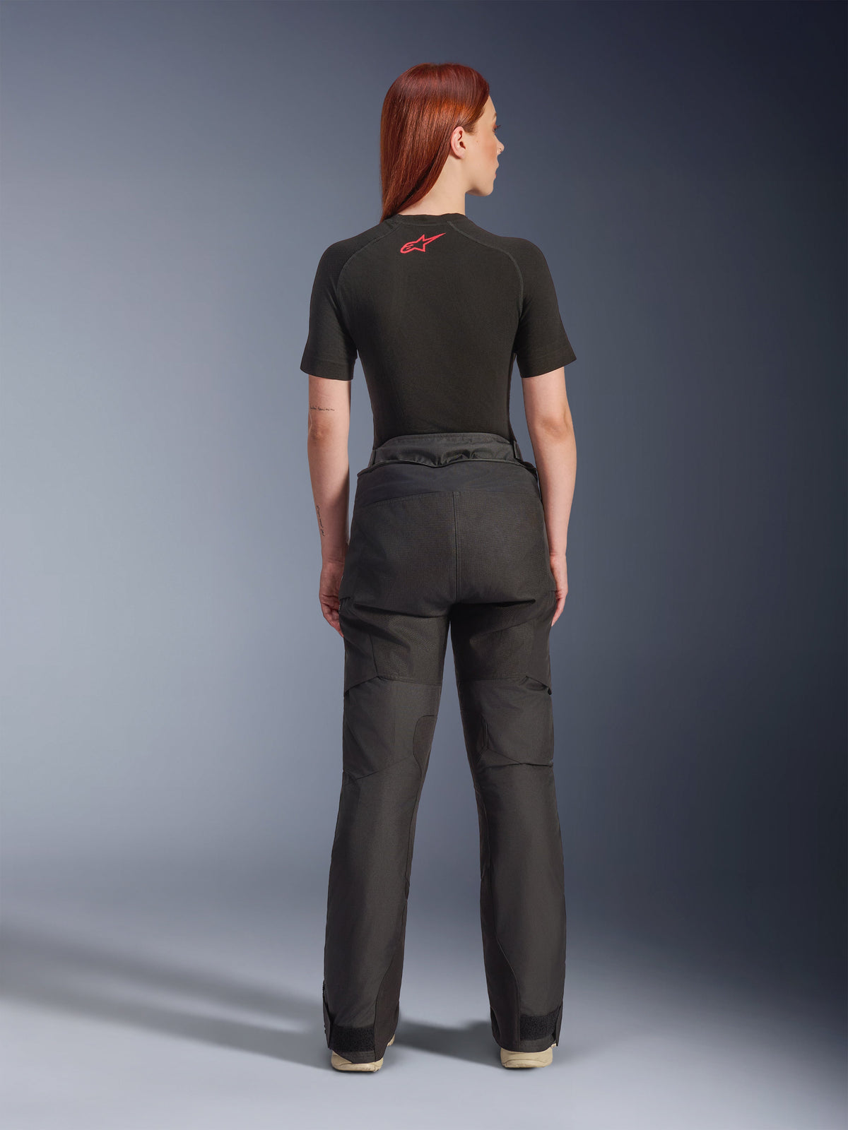 Rear view of a model wearing Alpinestars Stella Andes V4 Drystar® Pants in Black, paired with a black short-sleeve base layer featuring a red Alpinestars logo, adventure-touring motorcycle gear with durable textile construction.