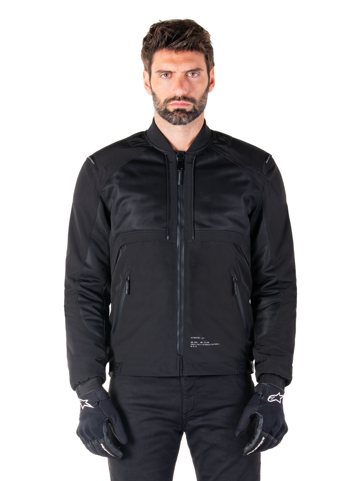Parka impermeable Alpinestars Mootant, chaqueta de moto negra, vista frontal en modelo masculino, con construcción textil modular con paneles de malla para ventilación, cuello acanalado y bolsillos con cremallera, usada con guantes negros con el logo de Alpinestars en blanco.