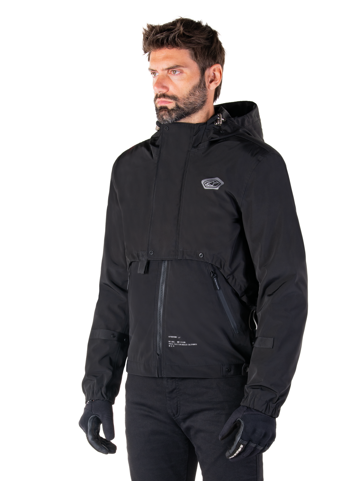 Alpinestars Mootant Waterproof Parka, chaqueta de moto modular negra, usada por un hombre con guantes y pantalones negros, cuenta con capucha, cremallera frontal y diseño utilitario por capas, estética urbana elegante con logotipo en el pecho.