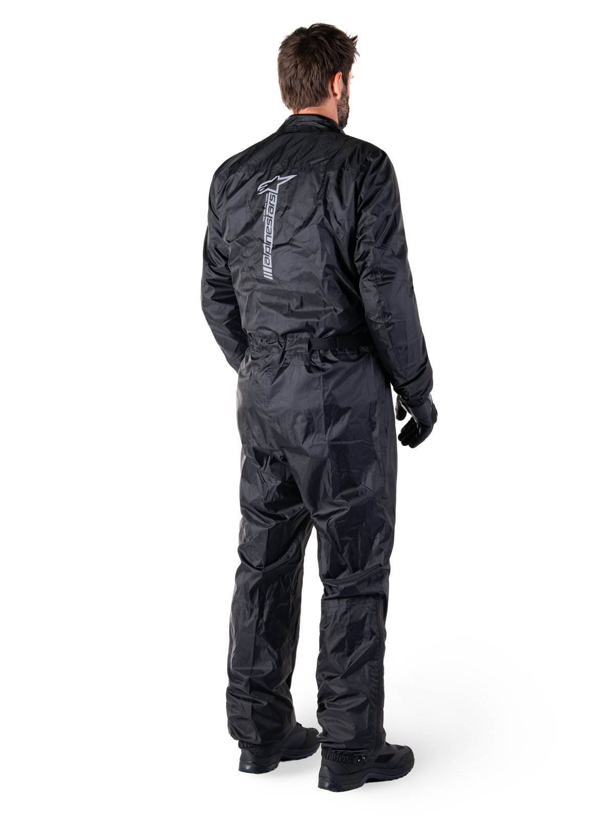 Alpinestars Hurricane Rain V2 Suit, mono de lluvia impermeable para motocicleta de una pieza en color negro, vista trasera con gran logotipo reflectante vertical, tejido ligero resistente al viento, usado con guantes y botas negras