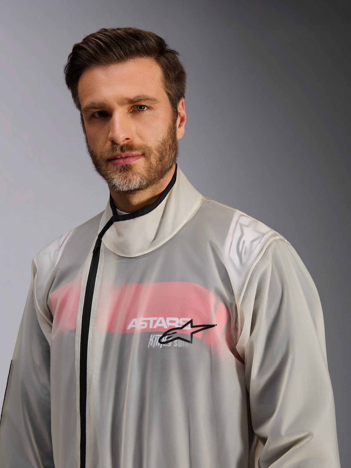Alpinestars Kart Rain Suit, sobre-mono impermeable de karting, transparente, lucido por un hombre, material impermeable transparente que muestra detalles de un mono de carreras rojo y blanco debajo, cuello alto con cremallera negra y logotipo de la estrella de Alpinestars en el pecho