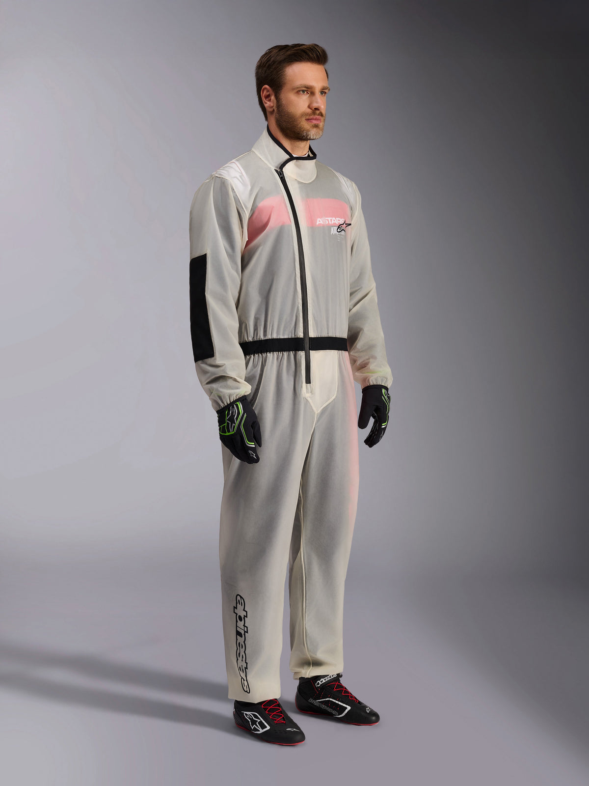 Alpinestars Kart Rain Suit, sobretraje de karting impermeable y transparente, material semitransparente que muestra un mono de carreras rosa y blanco debajo, con cremallera asimétrica negra, parche reforzado en el codo y banda negra en la cintura, usado con guantes de karting negros y calzado de competición