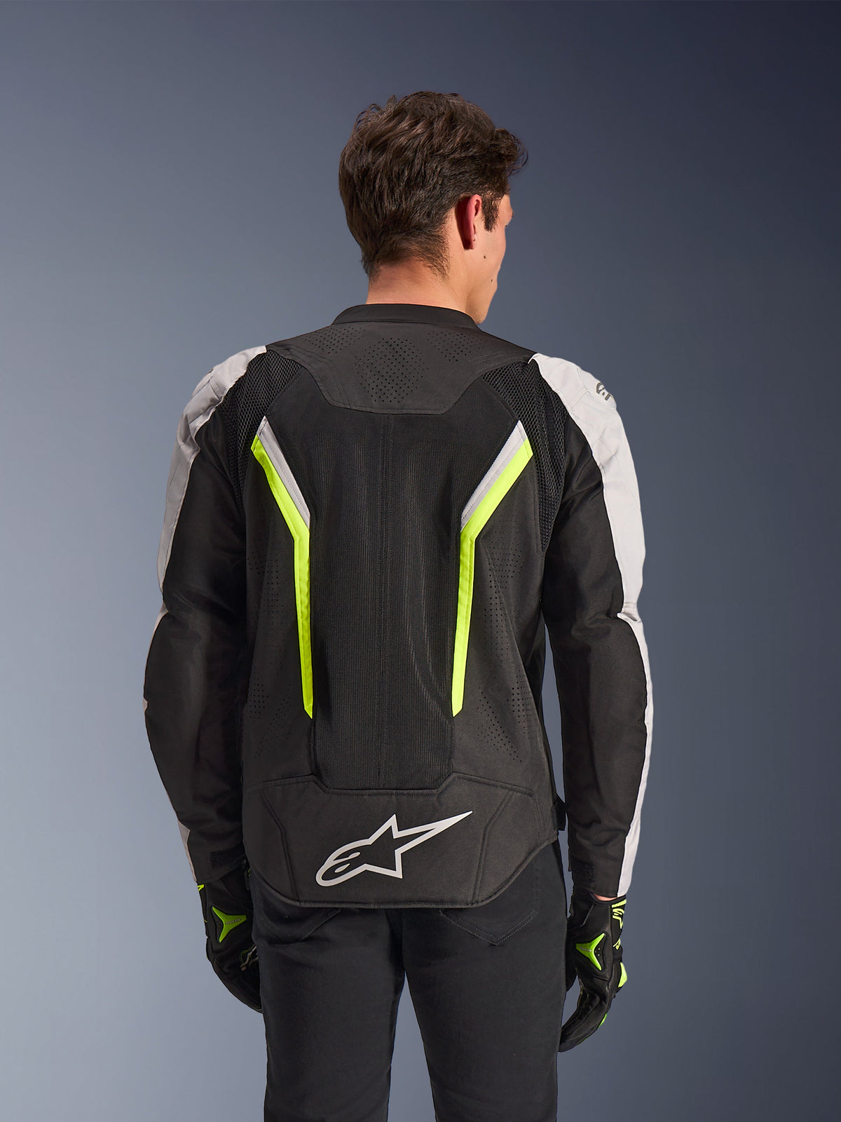 Chaqueta Alpinestars AST V3 Air, Negro Plata Amarillo Fluo, vista trasera en modelo, chaqueta textil para moto con paneles de ventilación de malla dual tech, franjas verticales en amarillo neón, mangas plateadas y logotipo blanco de Alpinestars en la parte inferior de la espalda.