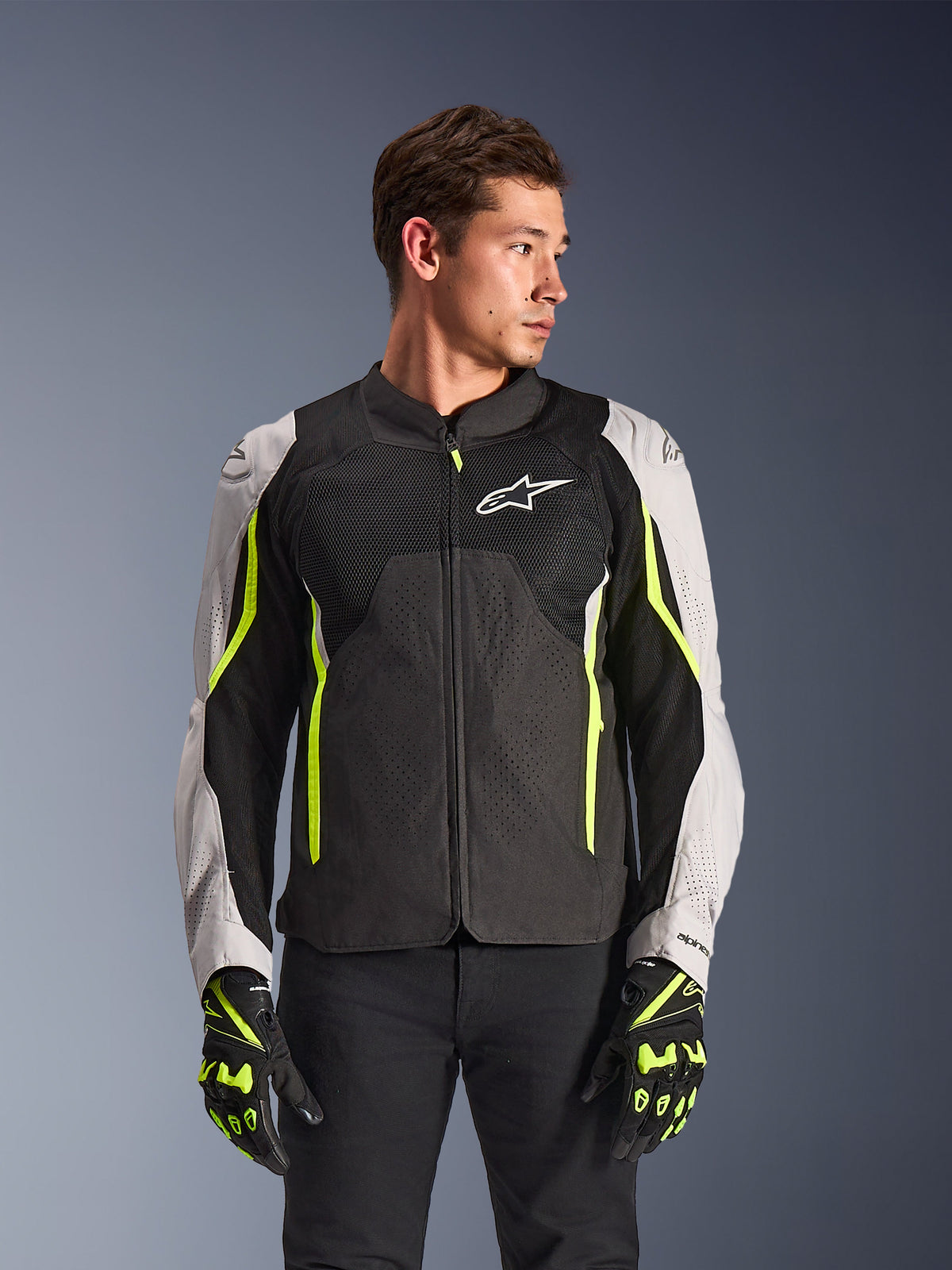 Alpinestars AST V3 Air Jacket, chaqueta de moto textil, negra y plateada con detalles en amarillo flúor, con grandes paneles de ventilación de malla y perforaciones láser, usada por un modelo con guantes a juego, equipamiento de conducción deportiva de alto rendimiento