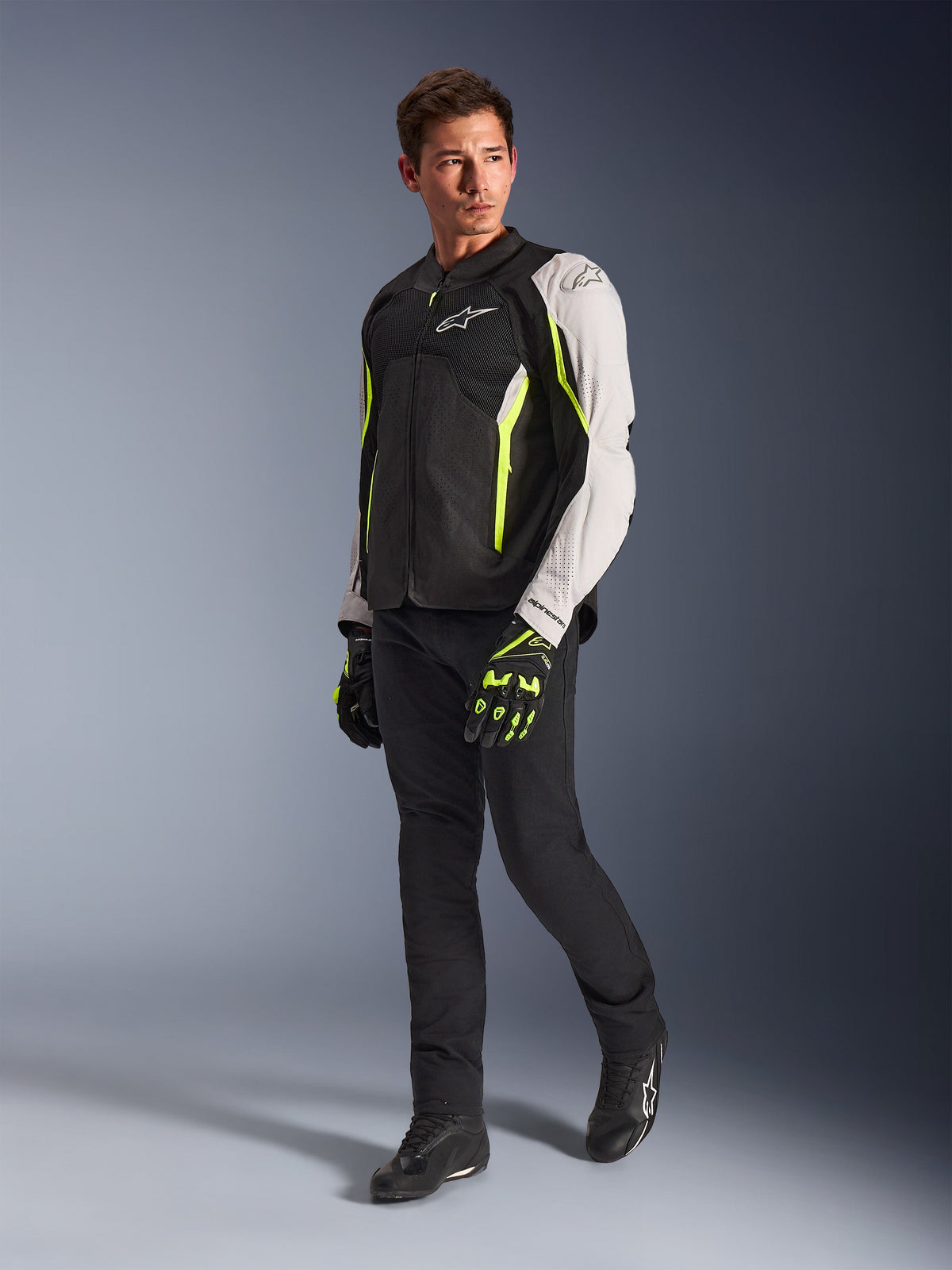 Alpinestars AST V3 Air Jacket, chaqueta de moto textil ligera en negro, plata y amarillo flúor, vestida por un modelo con pantalones negros y guantes a juego, con paneles de malla y perforaciones láser para conducción deportiva en climas cálidos.
